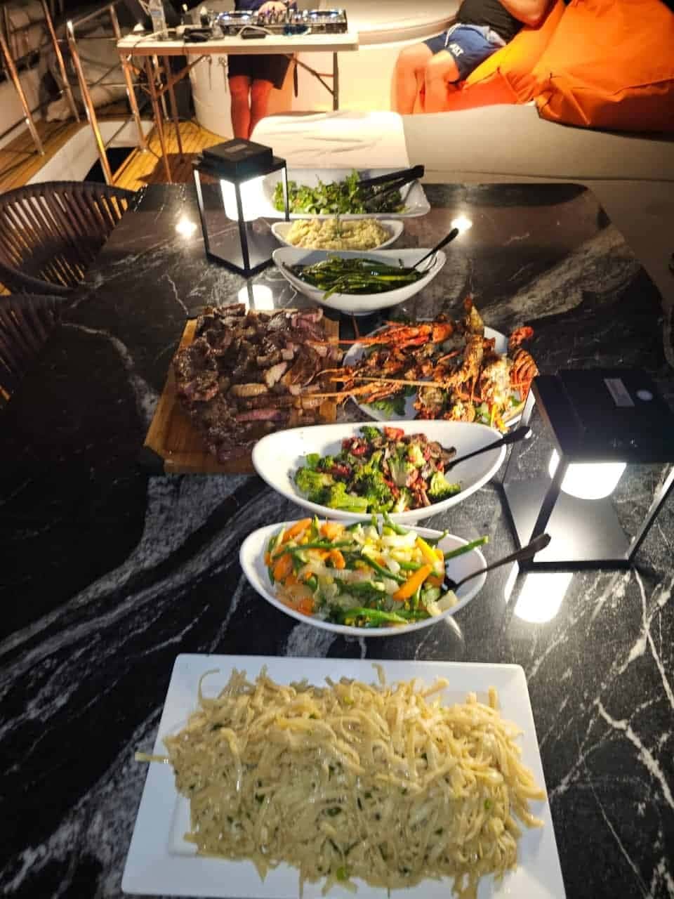 Grilled steak and lobster with salads and pasta laid out on a black marble table