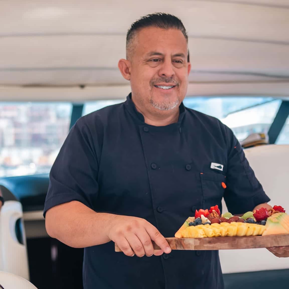 Lalo in a navy chef's jacket, carrying a board of fresh fruit
