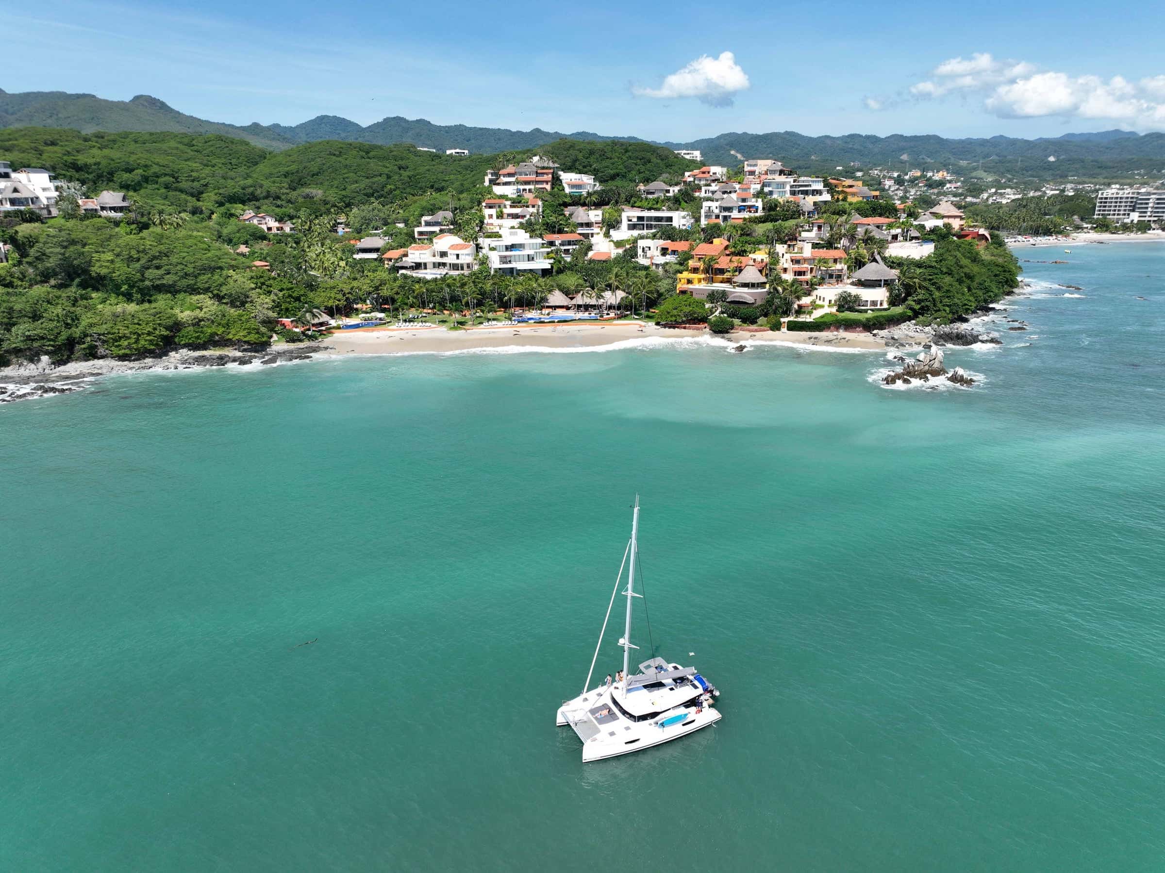Catamaran at anchor in turquoise water off a beach, below a hillside of houses and forest