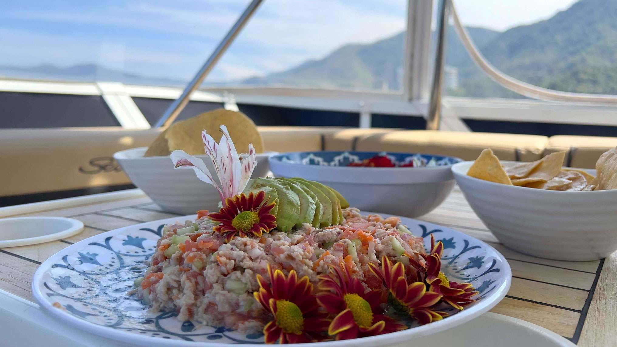 Plate of ceviche with avocado and flowers beside bowls of tortilla chips on a teak table