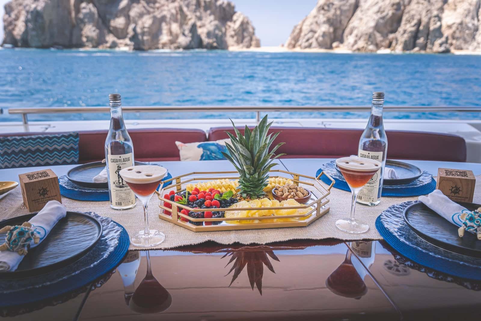 Aft deck table laid for four with a pineapple fruit platter, the rocks of Land's End behind