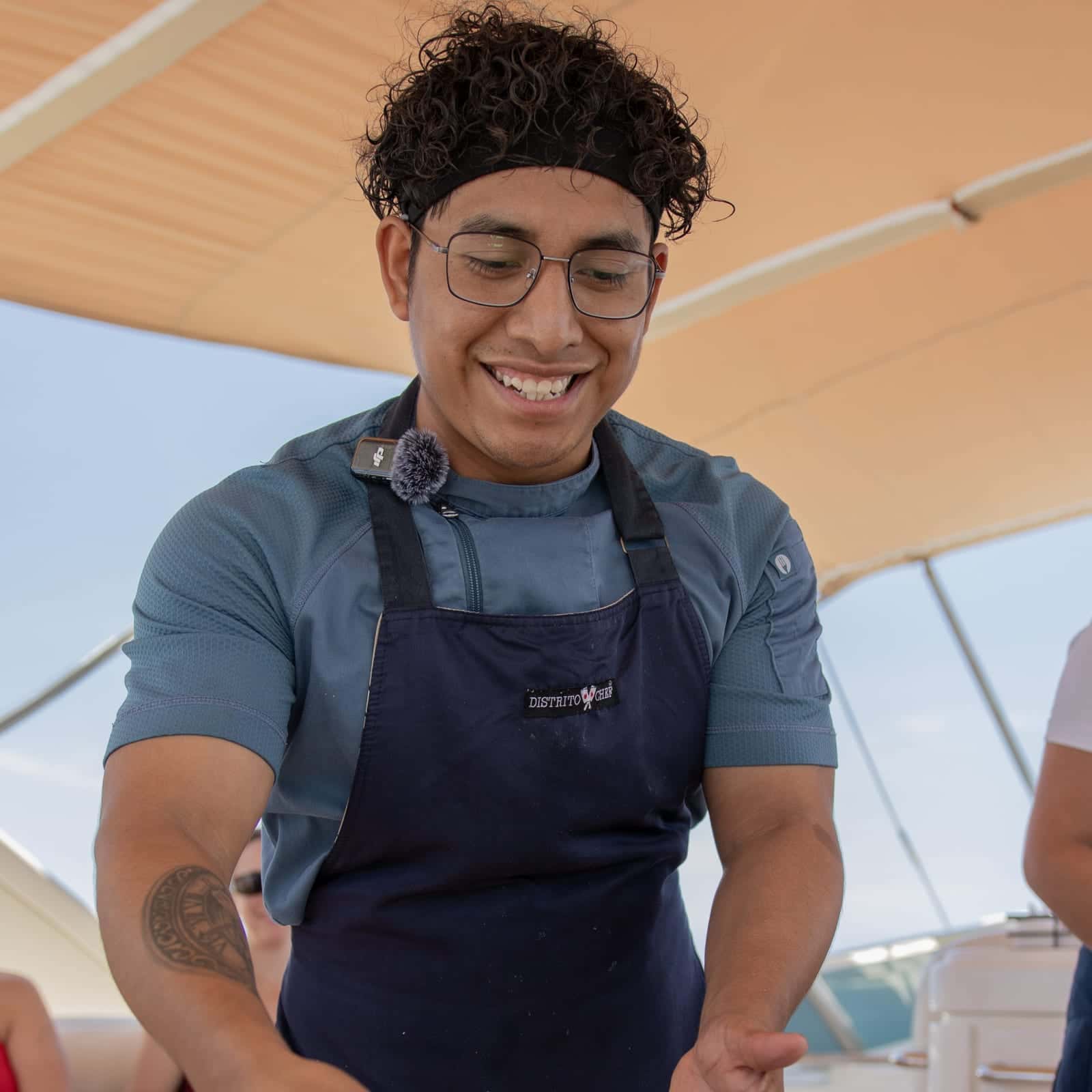 Santi smiling in glasses, a headband and a navy apron, under a deck's canvas shade