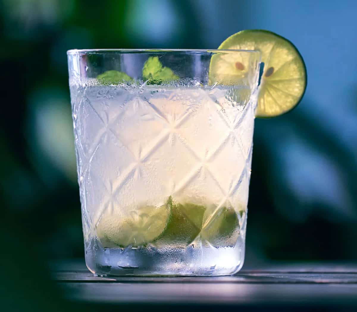 A Tater-ade in a cut-glass tumbler with mint and a wheel of lime, blue water behind