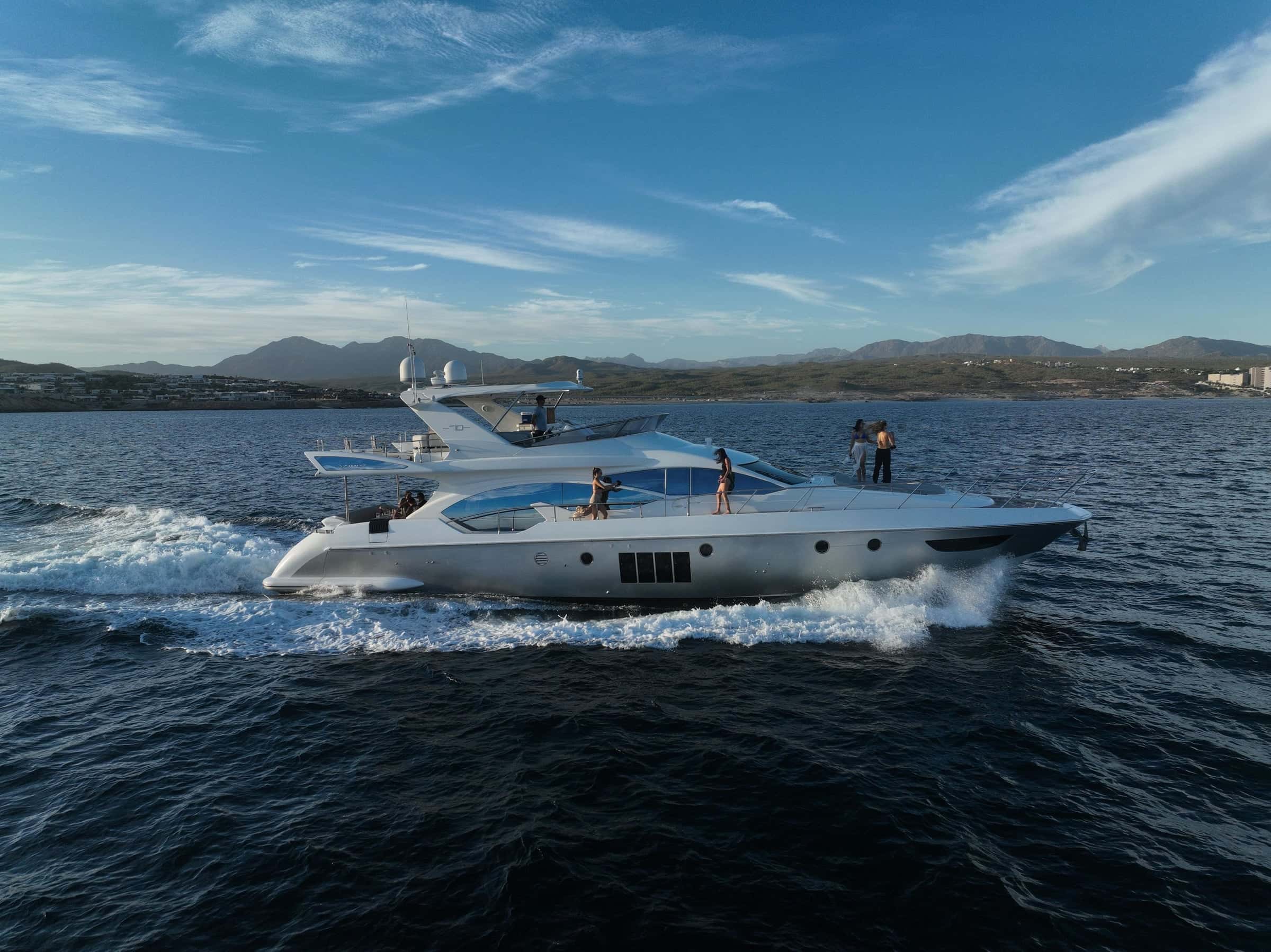The yacht under way with guests standing on the flybridge, spray along the hull and hills behind