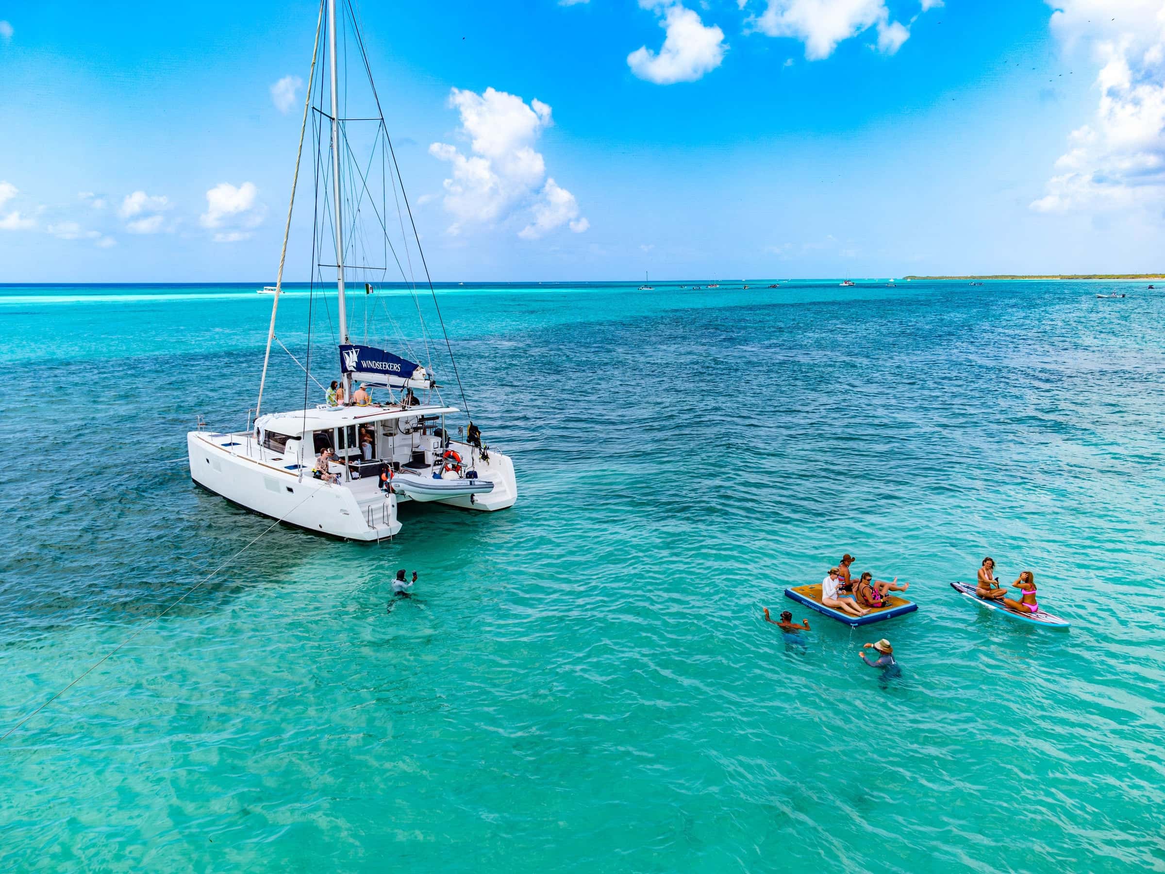 White sailing catamaran at anchor in turquoise shallows, guests swimming and floating beside her
