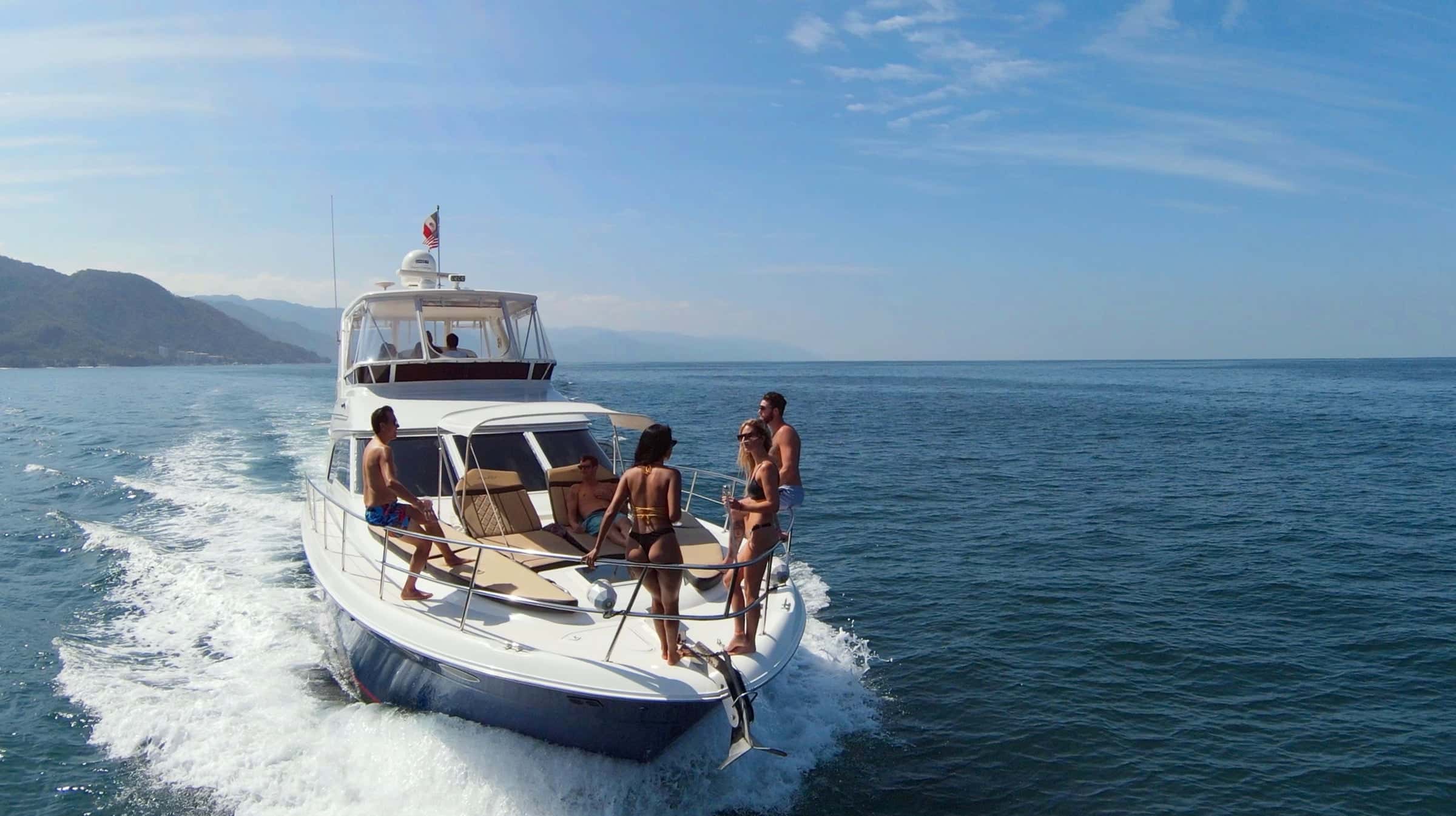 Motor yacht seen from astern and above at speed, guests on the swim platform and in the cockpit, green mountains behind