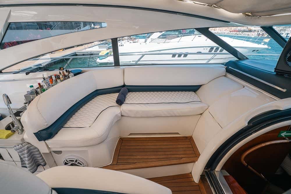 Cockpit lounge with a curved white sofa on a teak floor under the hardtop, bottles lined up at the bar