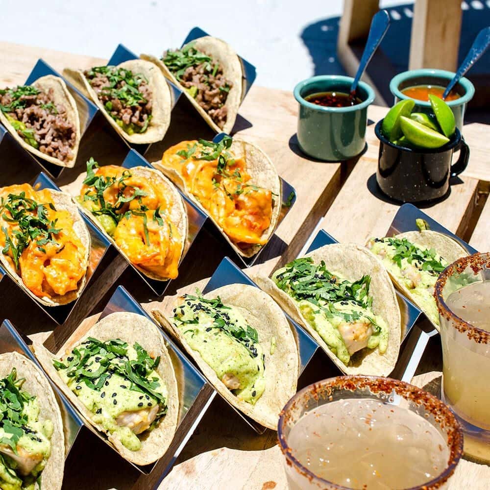 Tacos lined up in steel holders beside salsa and rimmed margaritas