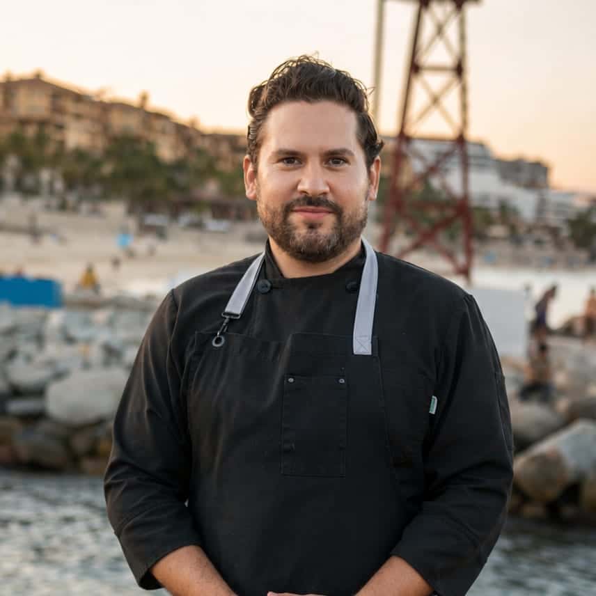 Christian in a black chef's jacket and apron on a rock jetty, a beach and hillside hotels behind