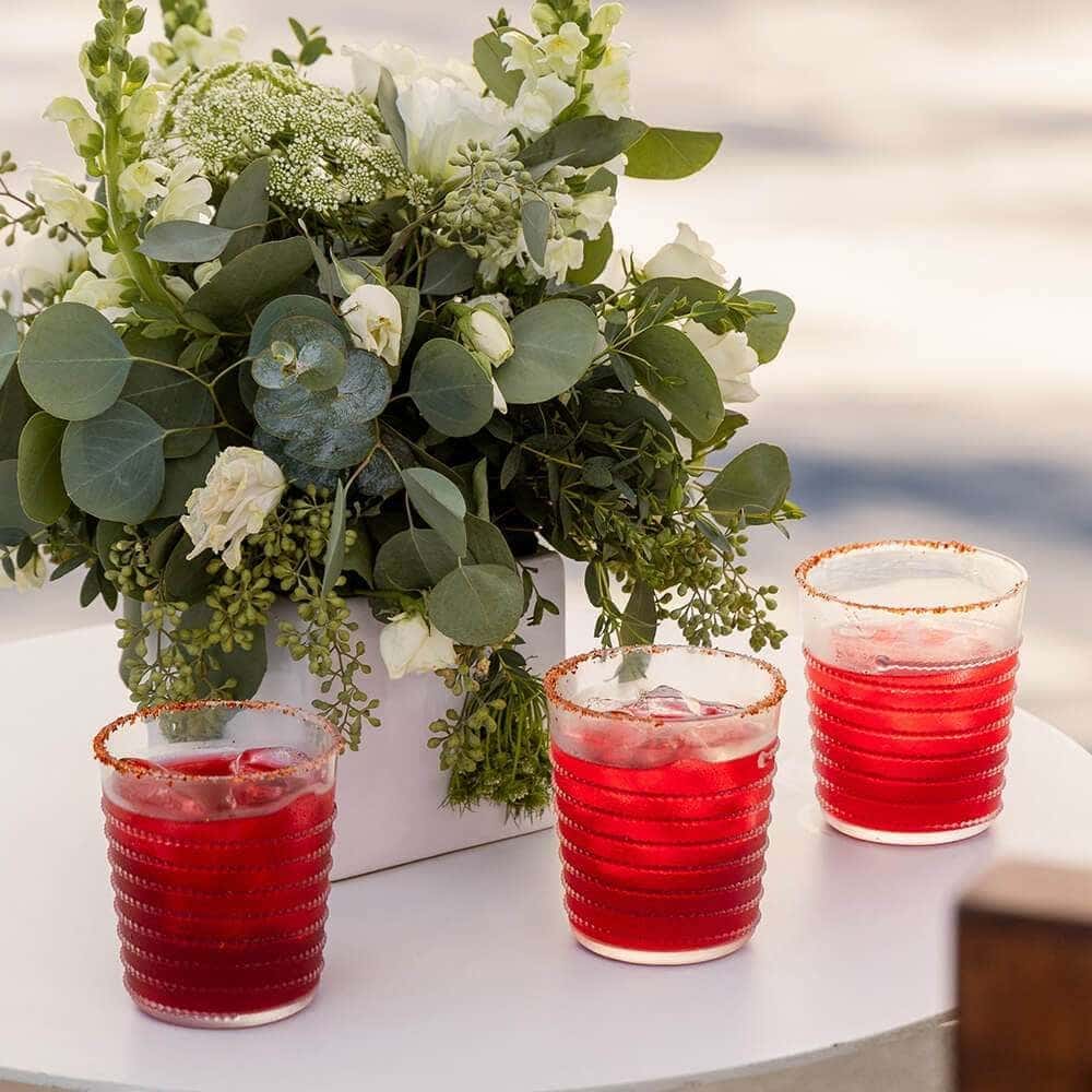 Three red cocktails with chile-salt rims beside a white flower arrangement