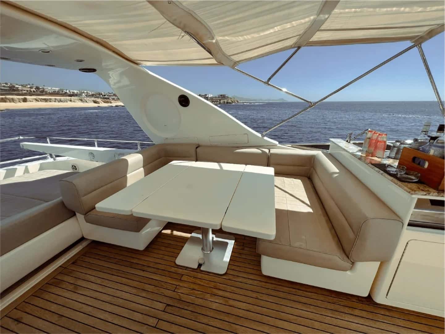 Flybridge under a bimini with an L-shaped settee, a white table and a bar counter, the coast beyond