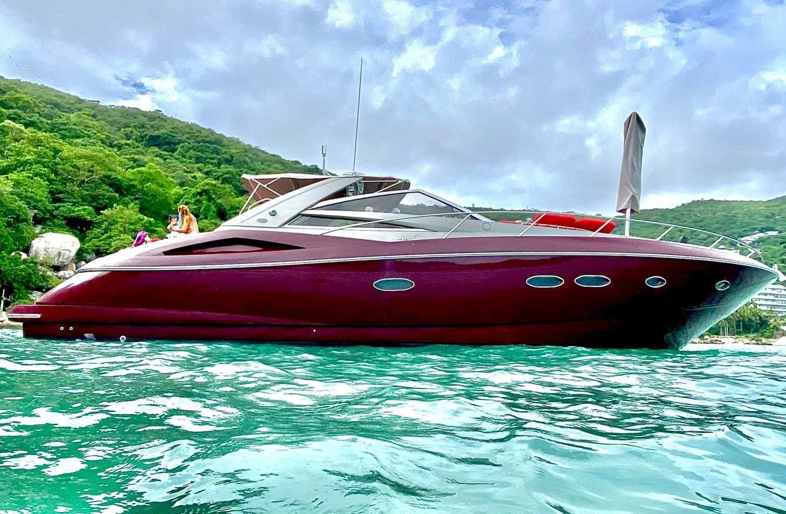 Dark red sports yacht in profile at anchor in green water, wooded hills behind