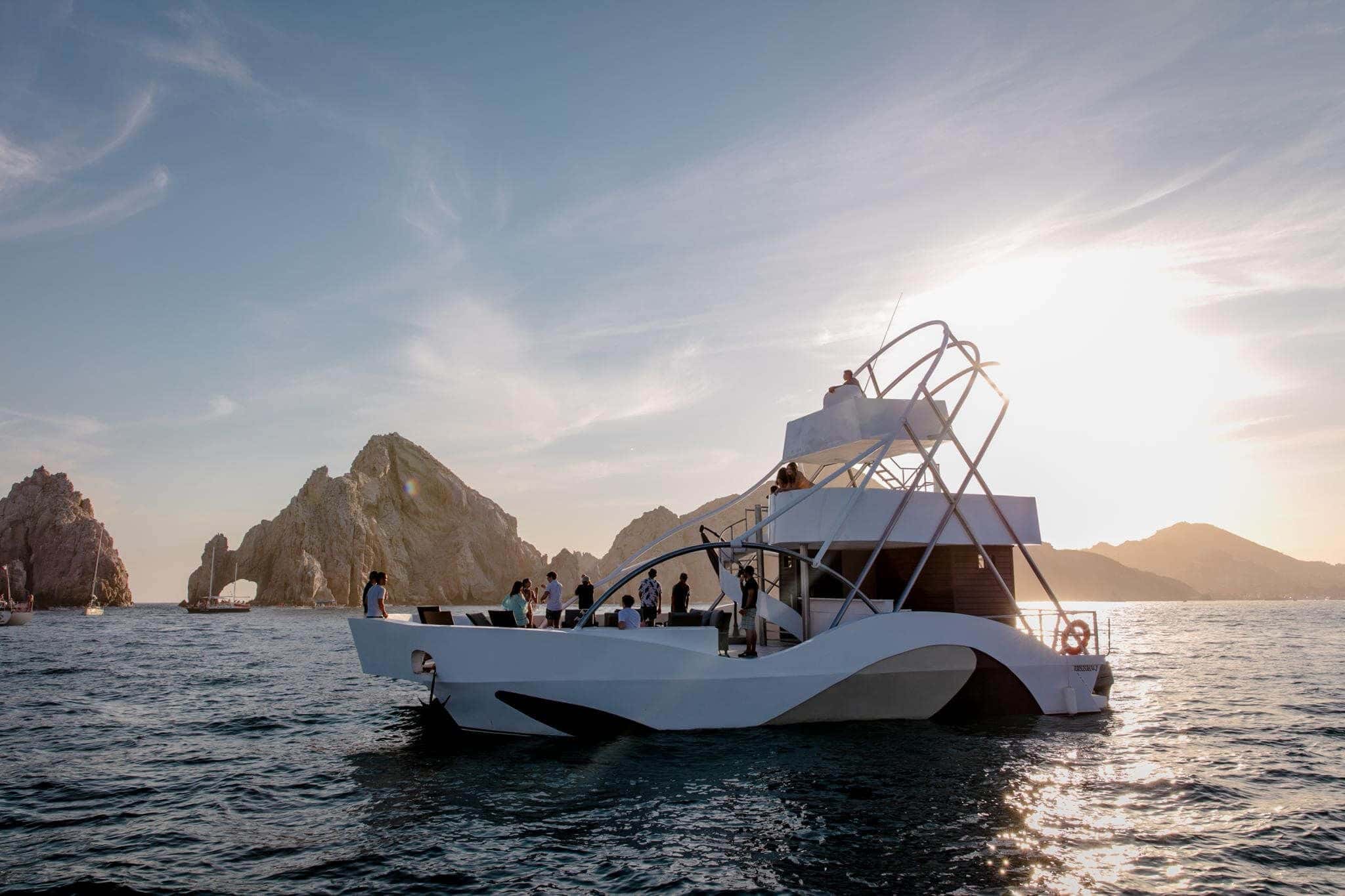 White catamaran with a curved upper deck and a lookout frame, the Arch rocks behind in late sun