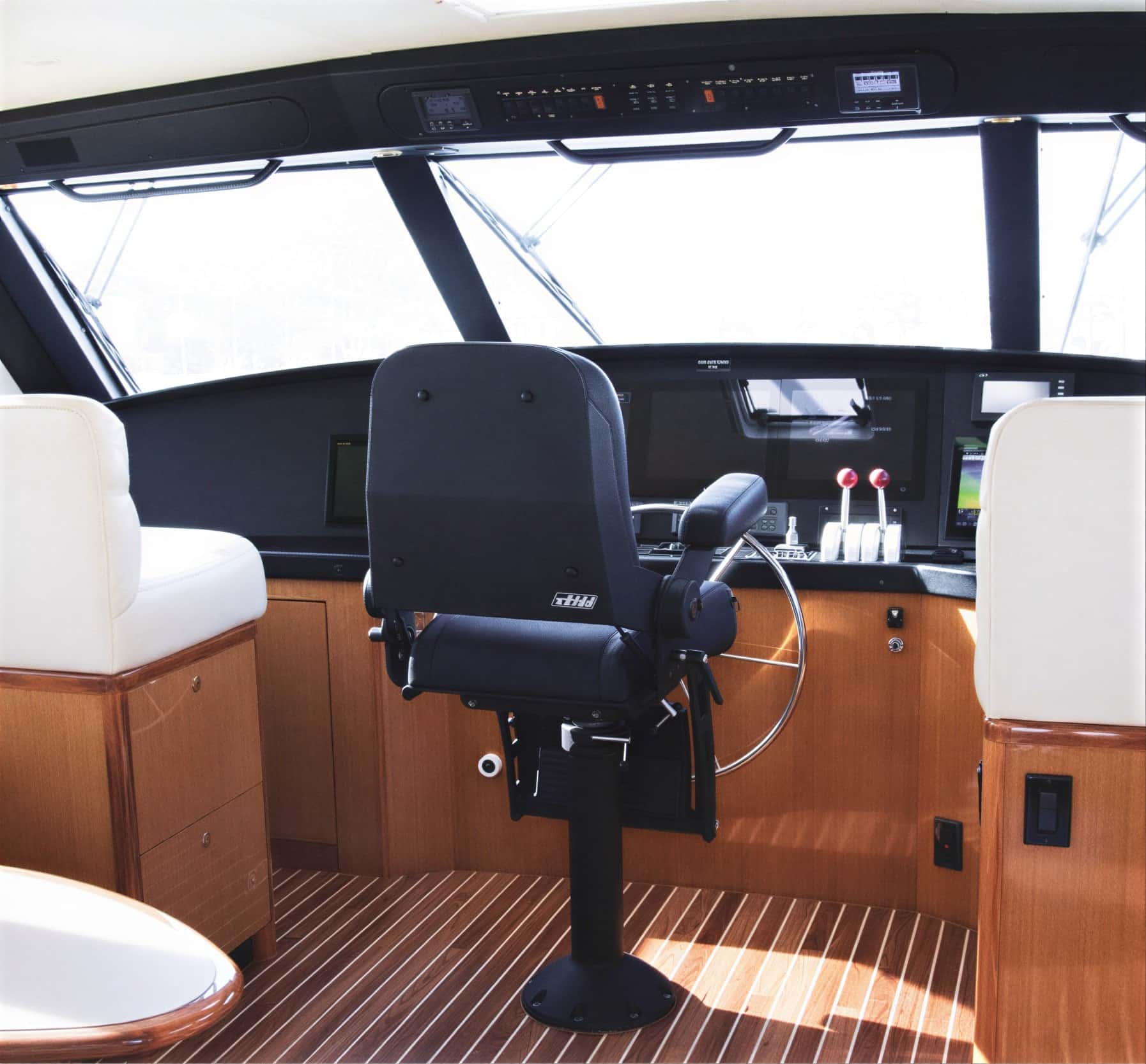 Helm in the enclosed bridge with a black captain's chair, wheel, throttles and screens, a teak floor and white seats either side