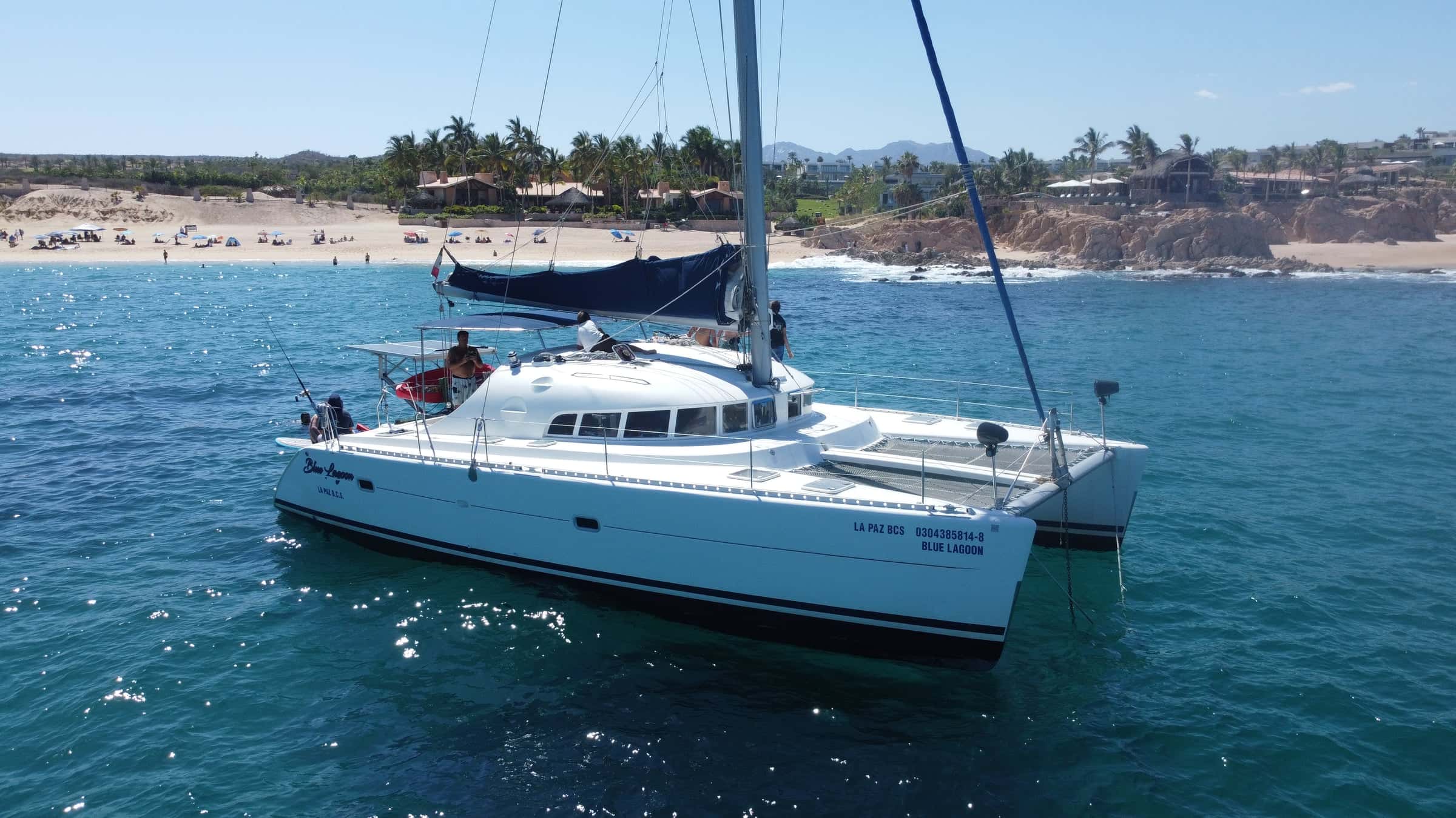 The catamaran anchored off a sandy beach with palms and low cliffs, guests on deck