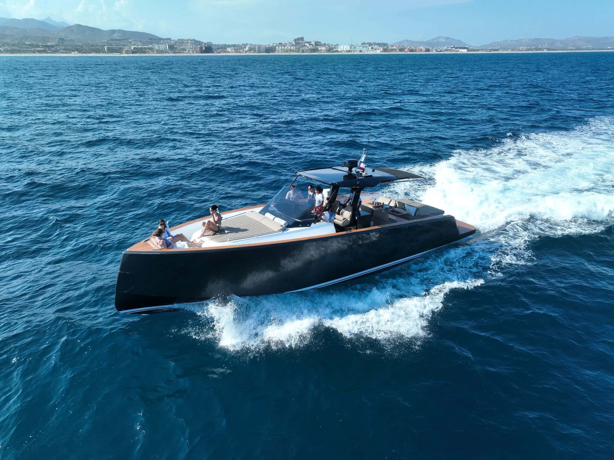 Aerial view of the dark-hulled Pardo running fast across deep blue water, a white wake behind and the coast beyond