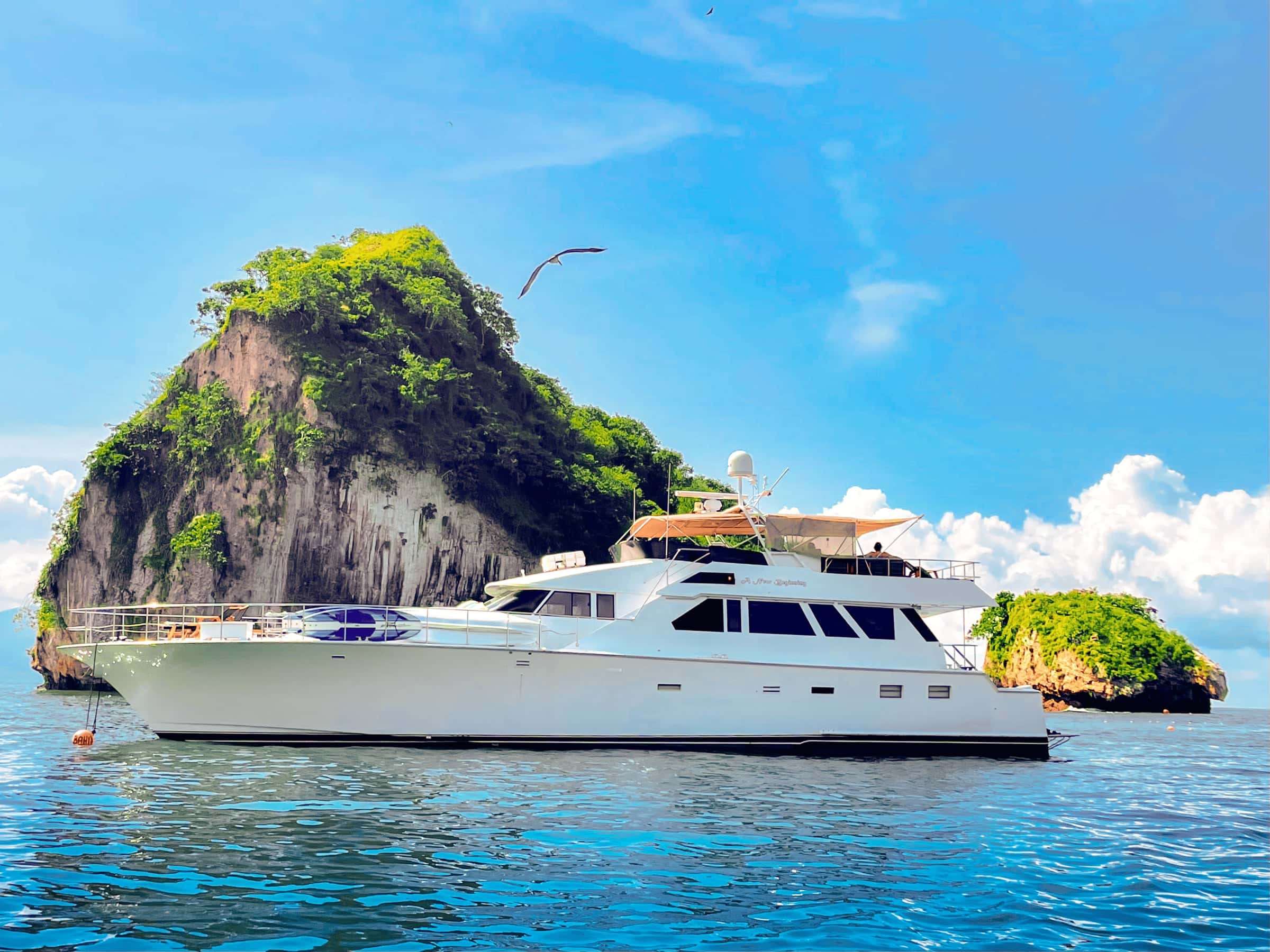 The yacht at anchor below a tall rock islet topped with green, on bright blue water