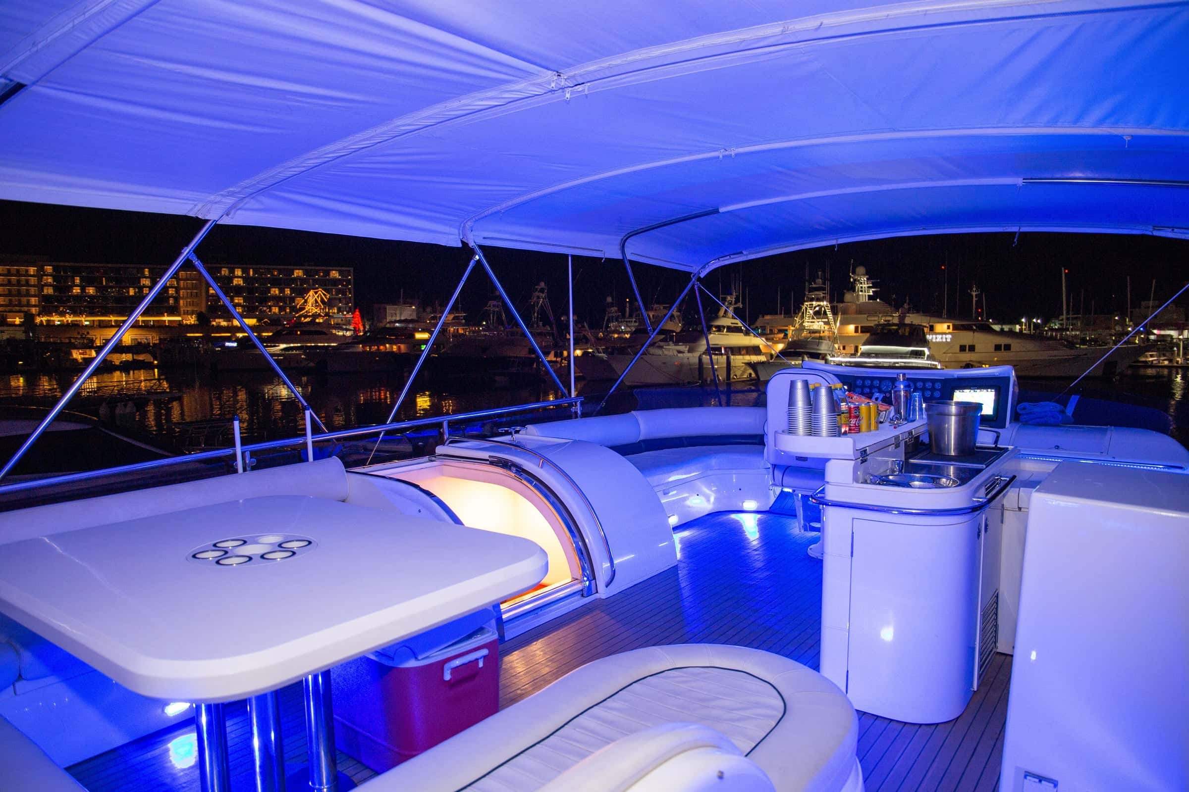 Flybridge at night in blue light, the wet bar with bottles and an ice box beside the table, the lit marina beyond