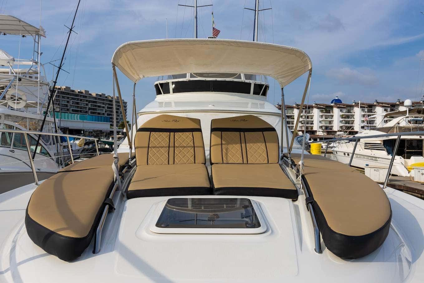 Two tan sun loungers and a cushioned bench on the foredeck under a canvas Bimini, the marina behind