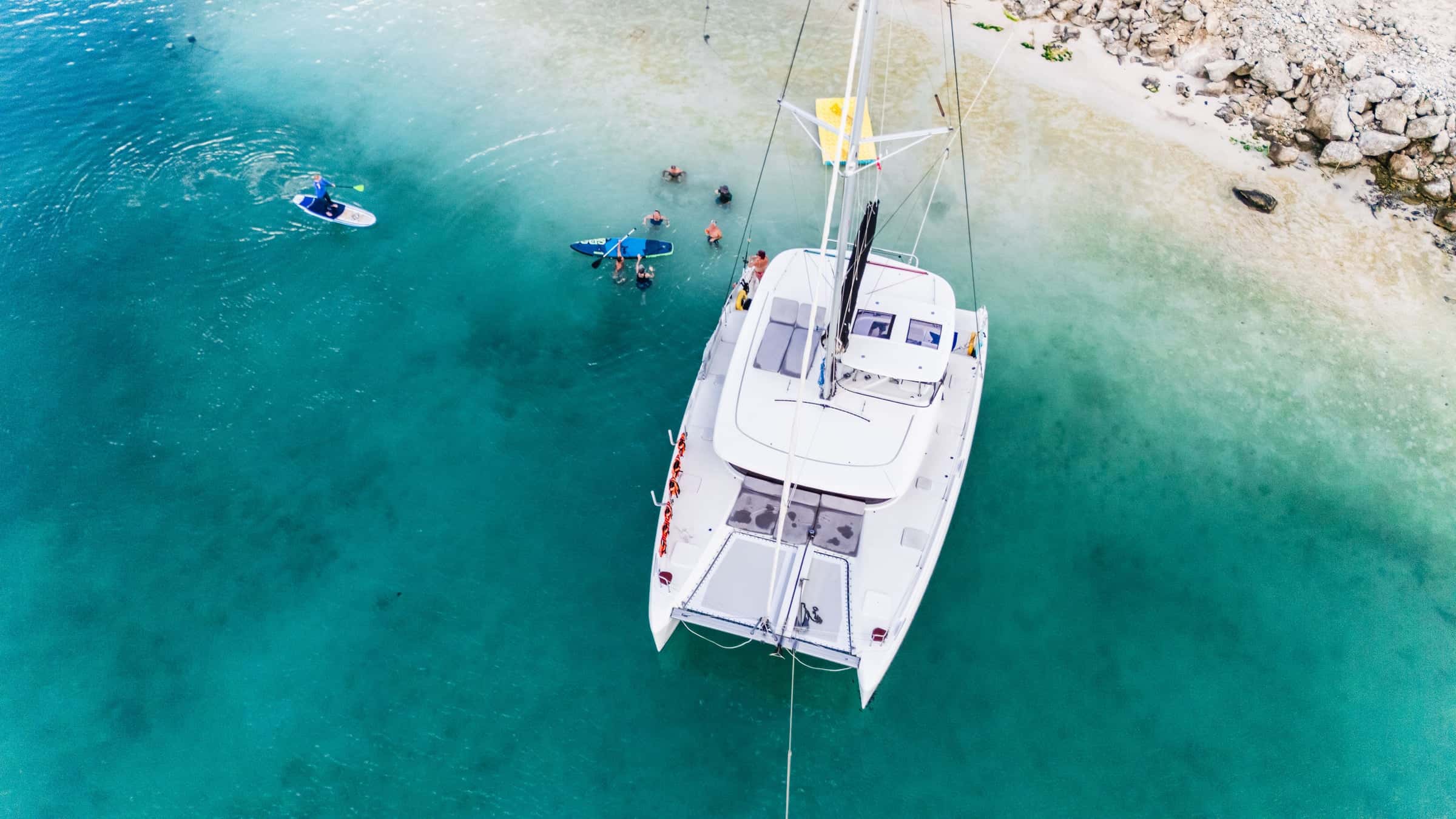 Catamaran from above, anchored in shallow turquoise water off a white beach, a paddleboarder and swimmers nearby