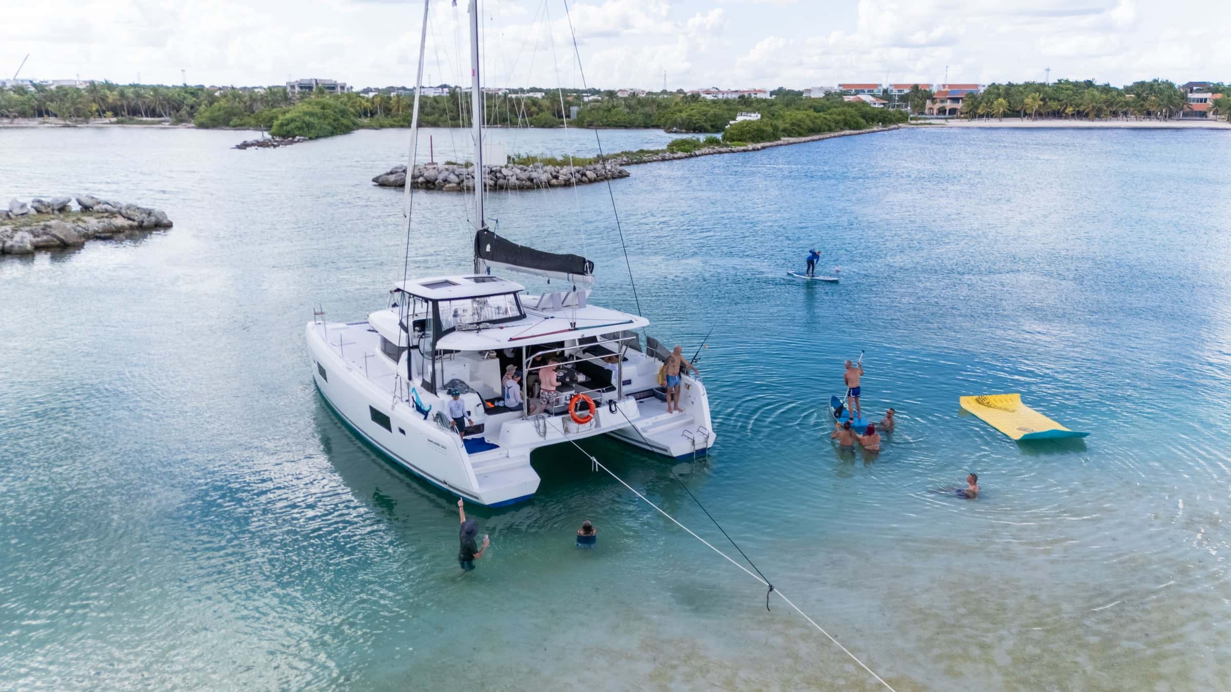 Catamaran anchored in a calm lagoon, guests swimming off the stern beside a yellow floating mat and a paddleboarder