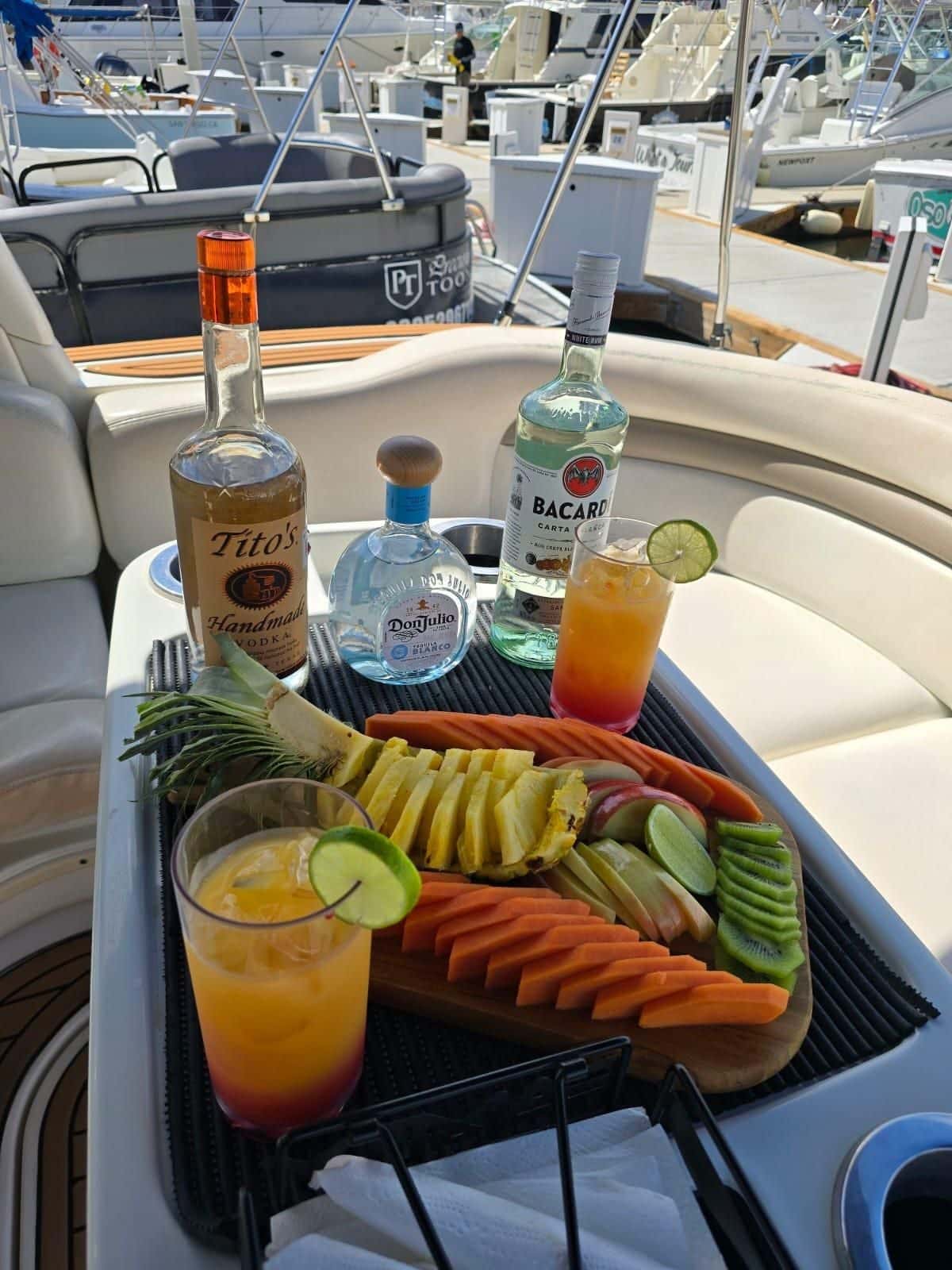 A board of sliced pineapple, papaya and cucumber with mixed drinks and bottles of tequila, rum and vodka on the cockpit table