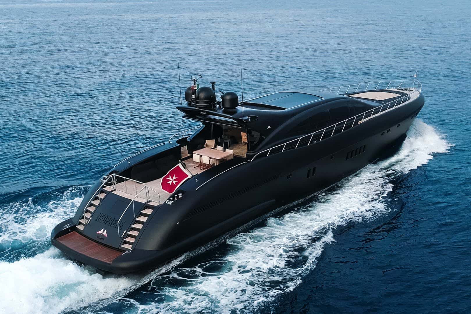 Matte black yacht under way seen from above and astern, a table and chairs on the aft deck and a Maltese flag at the stern