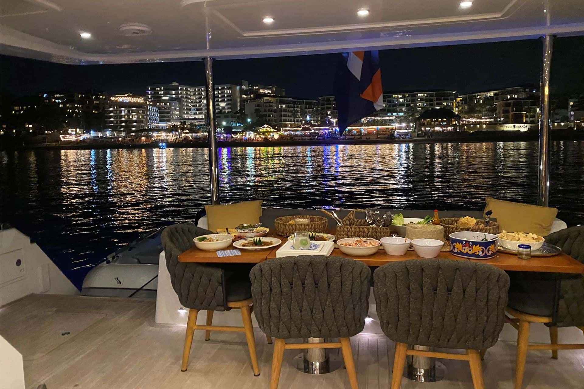 Dinner dishes laid out on the aft deck table at night, woven chairs and the lit marina beyond