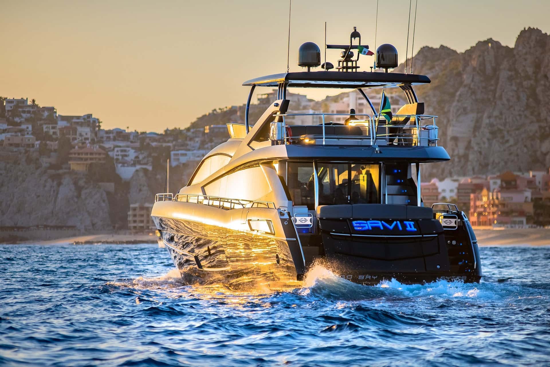 The yacht under way at sunrise seen from the stern quarter, the name lit in blue and the hillside behind