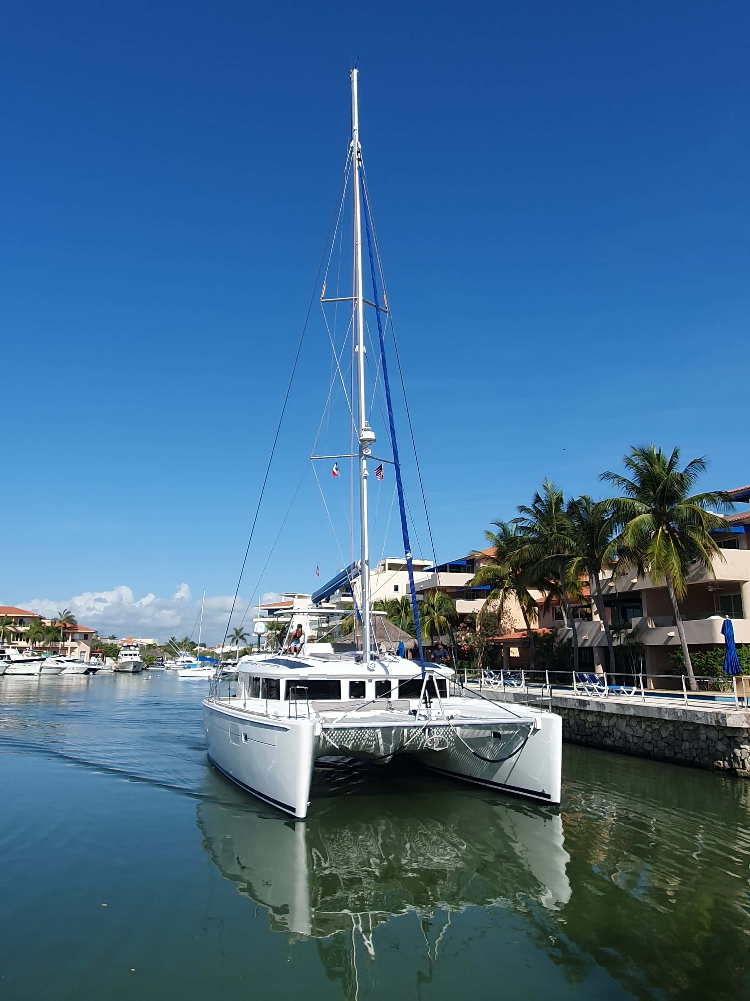 The catamaran bow-on in the calm marina channel, palms and houses along the bank