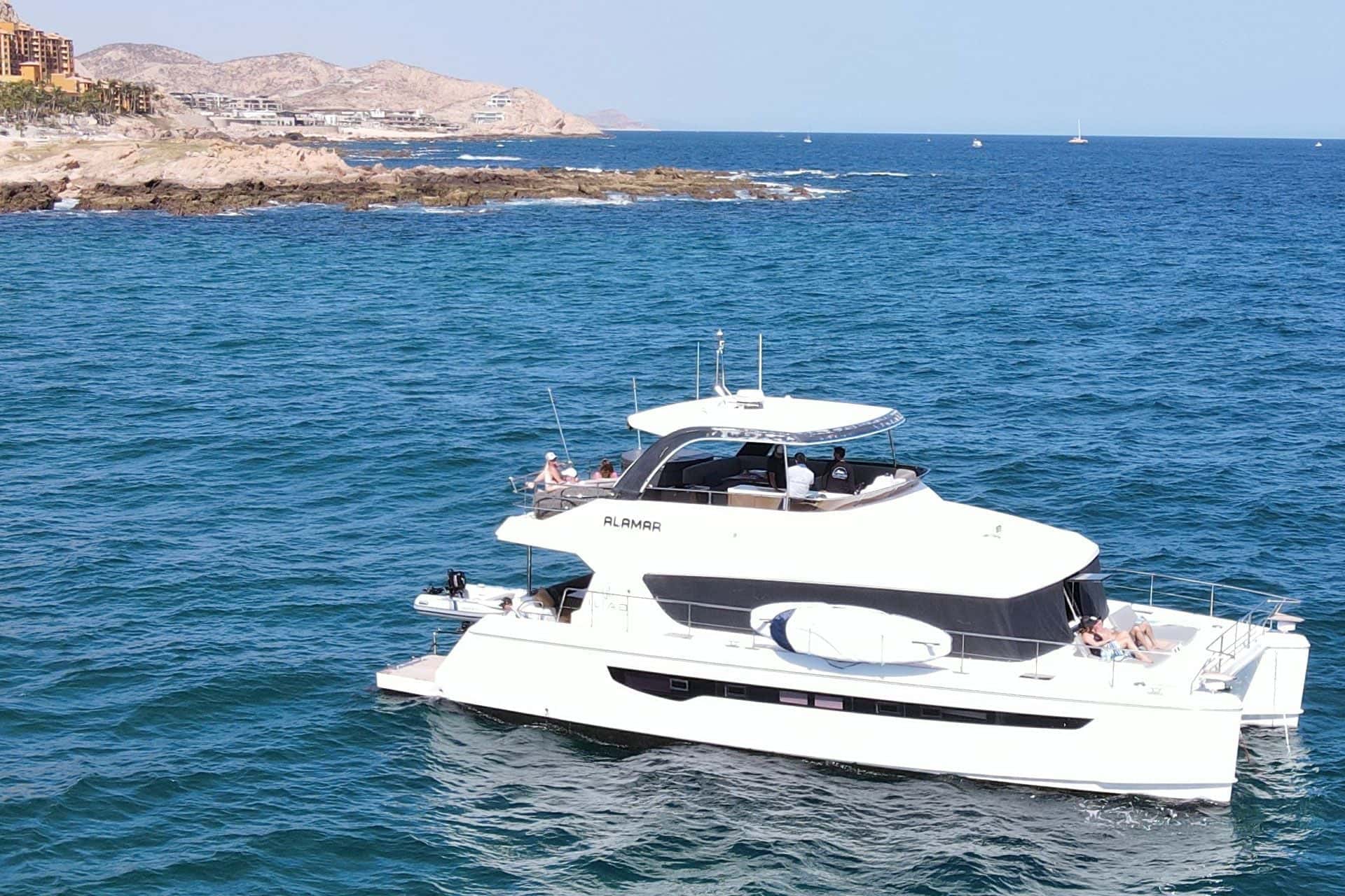 Power catamaran from above on deep blue water off a rocky point, guests on the foredeck
