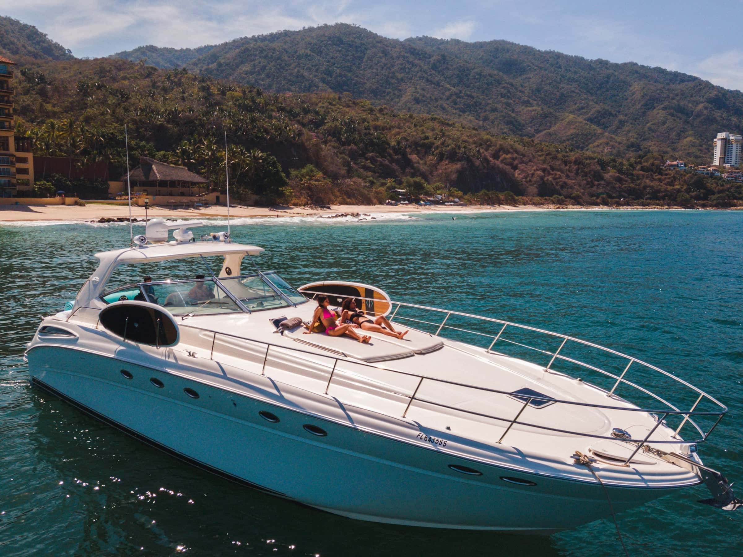 Express motor yacht seen from above the bow, two guests on the foredeck beside a paddleboard, a palm-backed beach and mountains behind