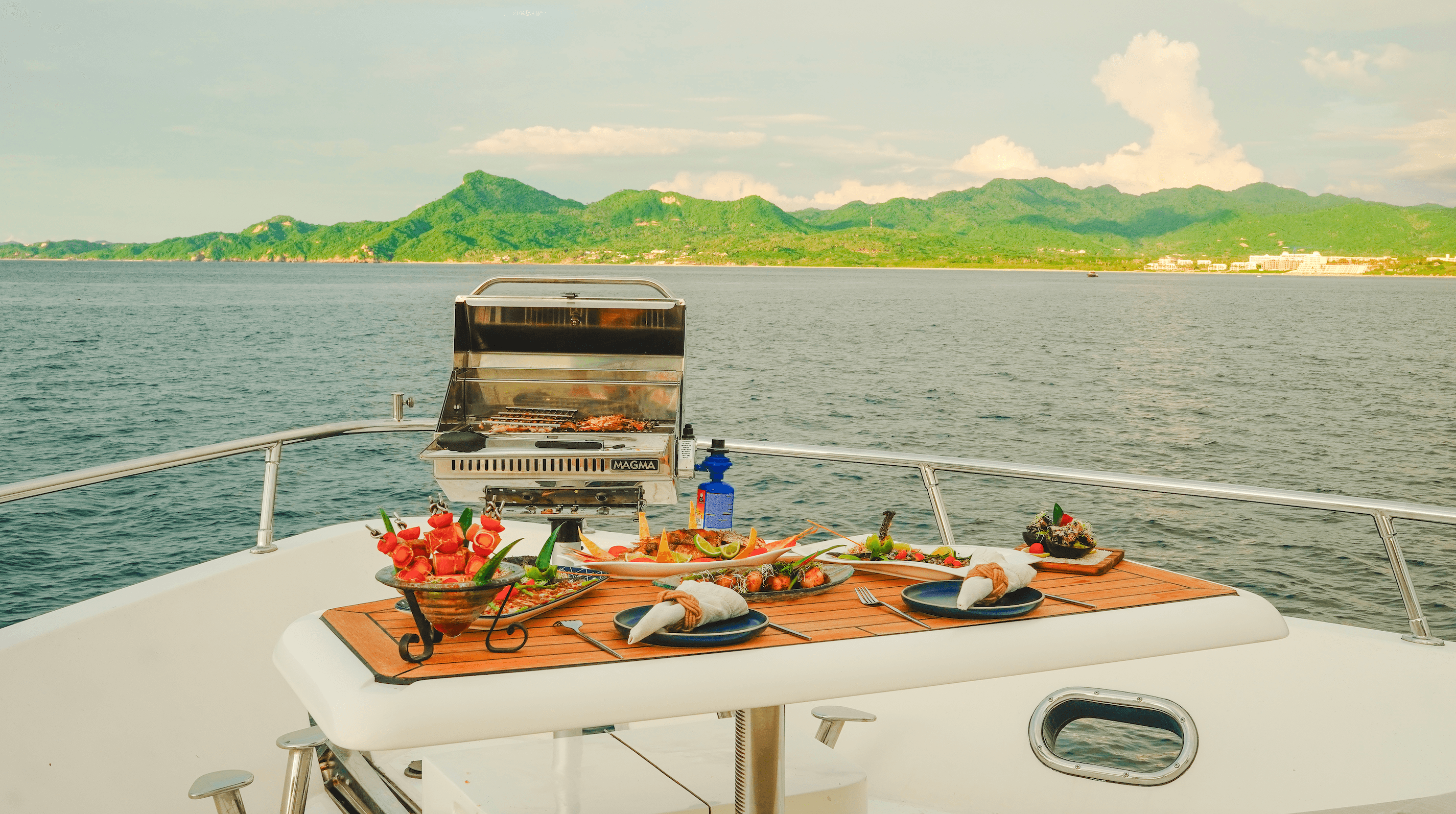 Food spread on the teak table at the bow beside the grill, with the hills of Banderas Bay behind