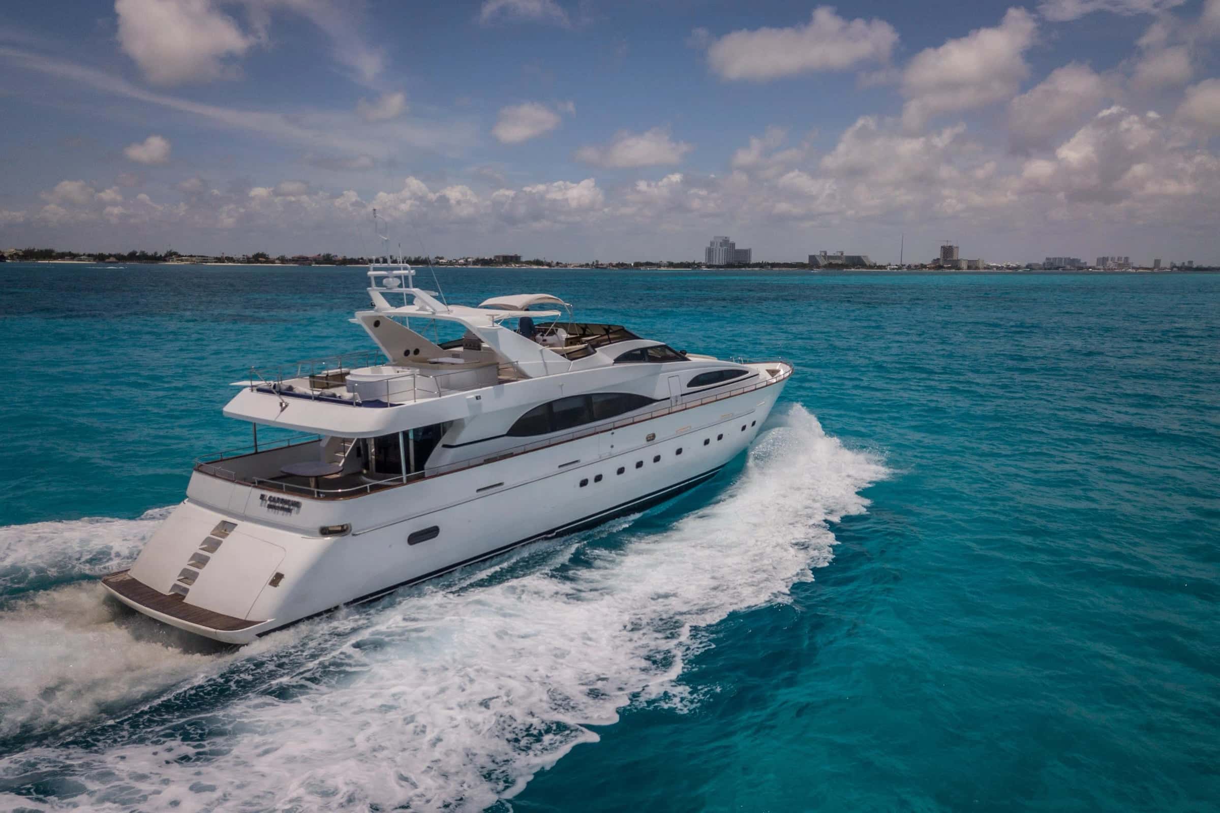 The yacht under way at speed from the bow quarter, a white wake on blue water and the Cancún shore behind