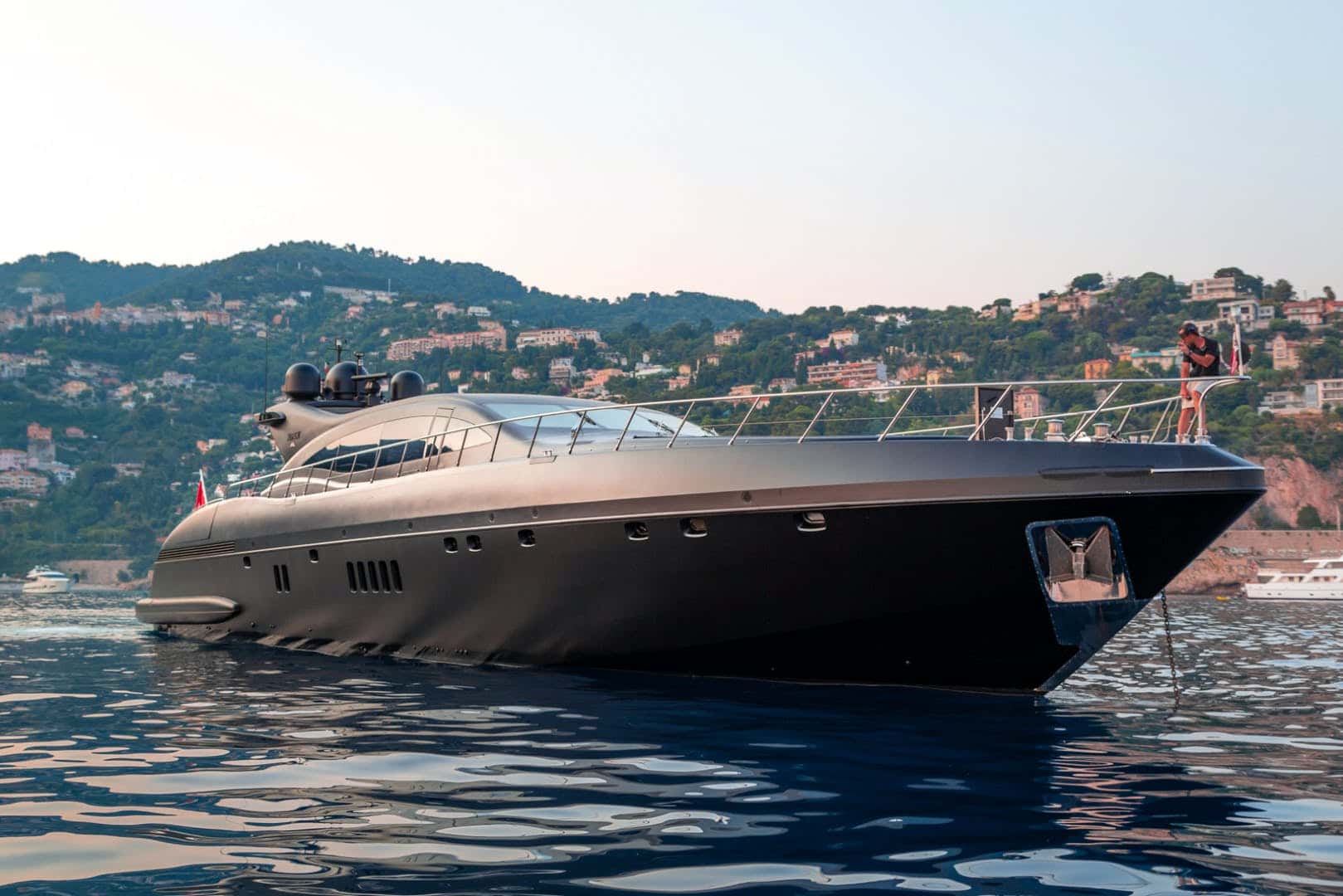 Matte black yacht at anchor, a crew member on the bow and a green hillside with villas behind