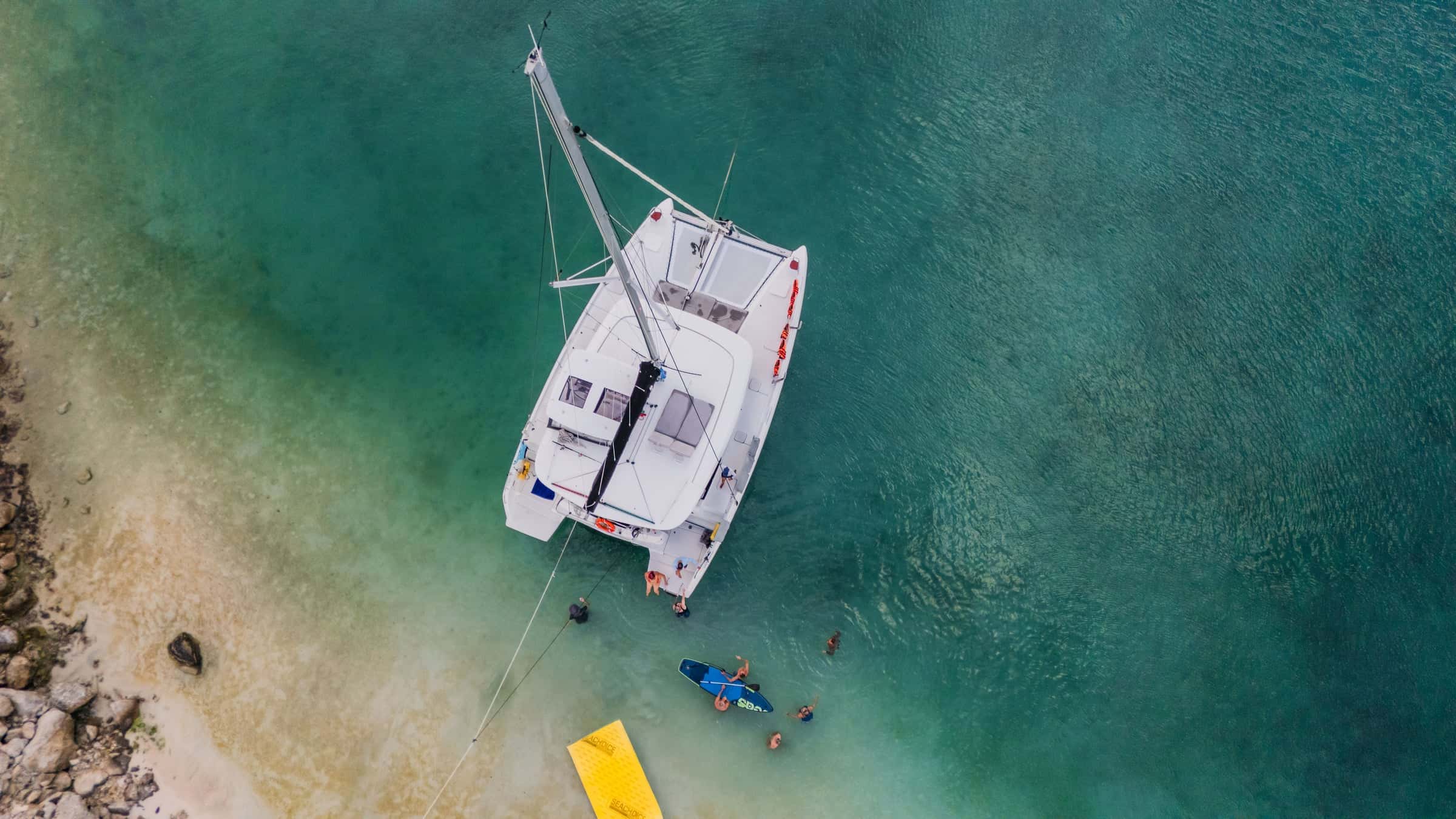 Top-down view of the catamaran at anchor off a sandy shore, swimmers and a yellow floating mat at the stern