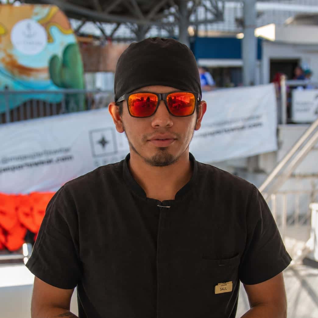 Saul in a black chef's top, a cap and orange sunglasses at the dock