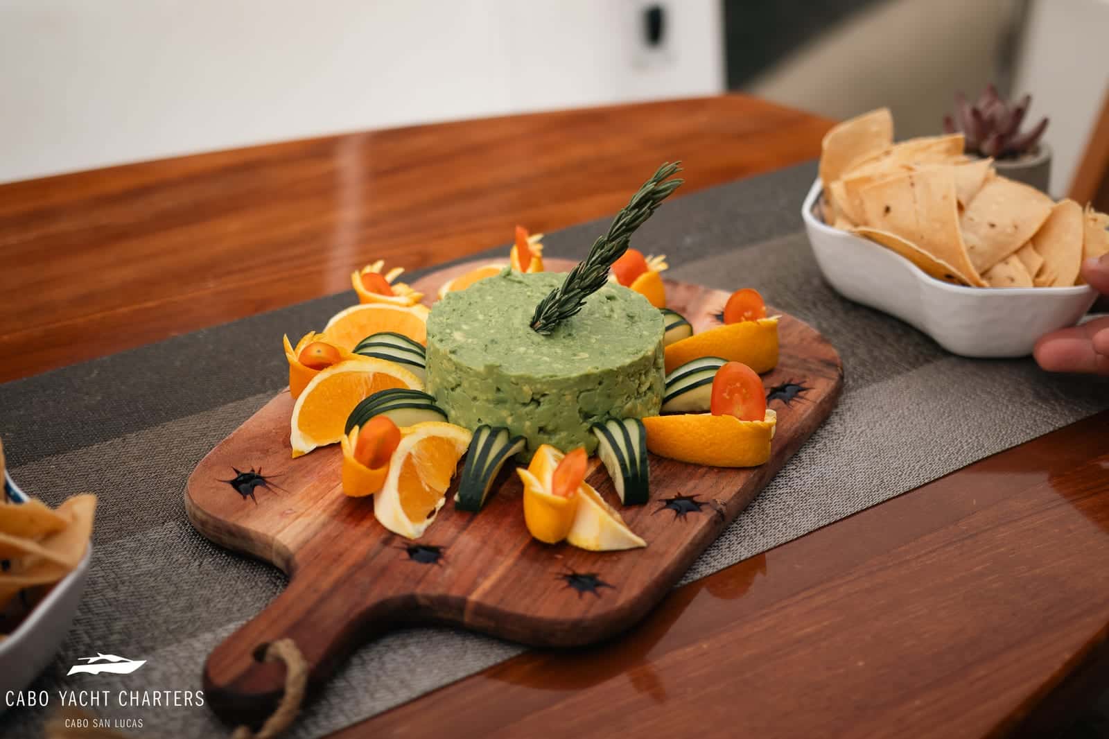 Guacamole on a wooden board ringed with orange slices, cucumber and cherry tomatoes, a bowl of chips beside it