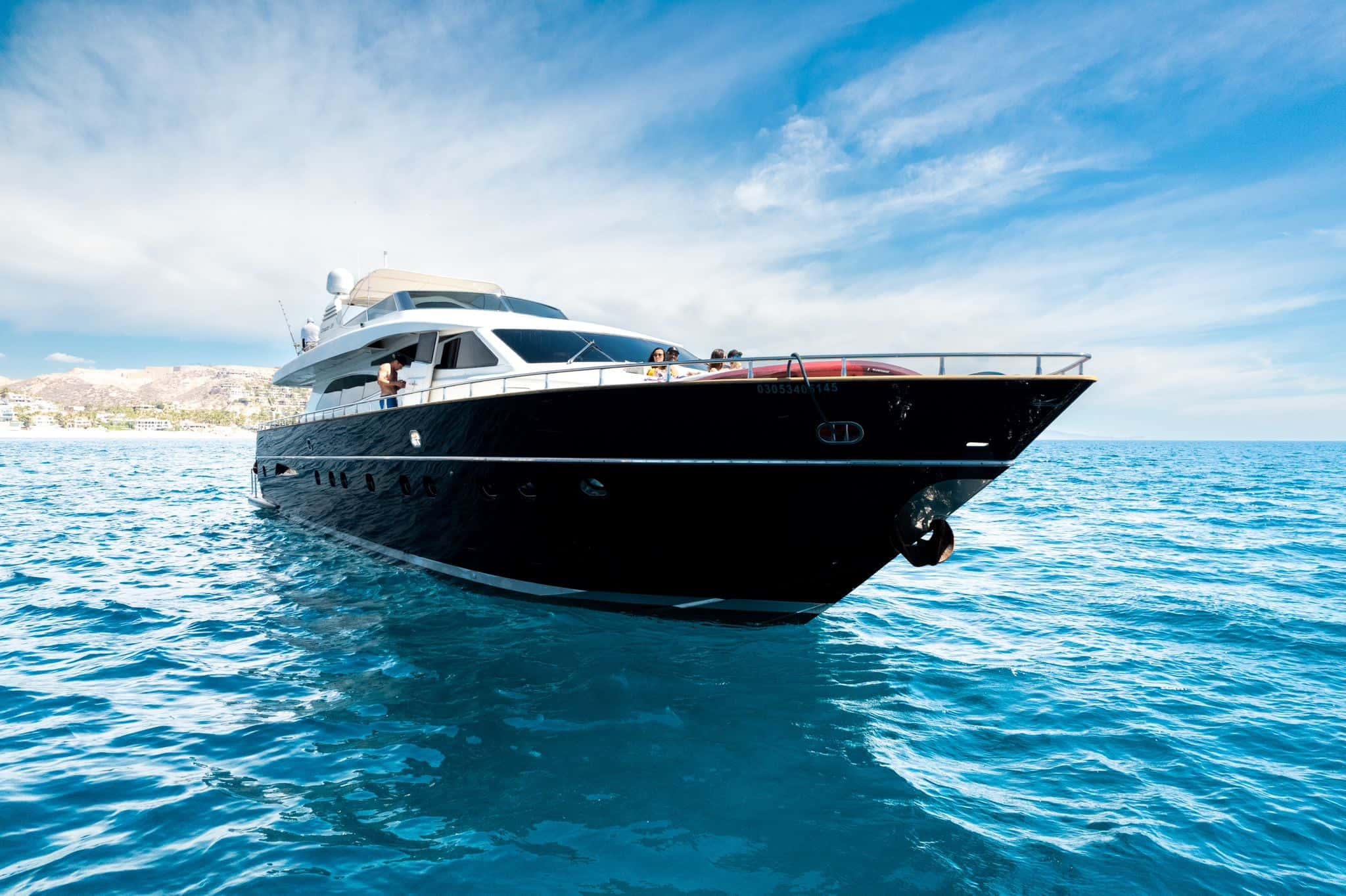 Black-hulled motor yacht at anchor seen from the bow quarter, flybridge above and the Cabo coast behind