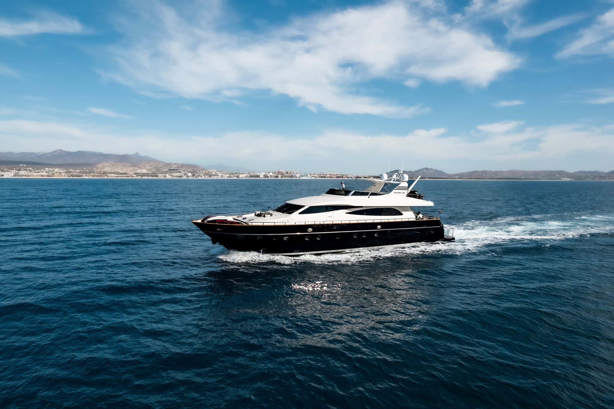 Black-hulled motor yacht under way with a white wake, the coast and hills behind