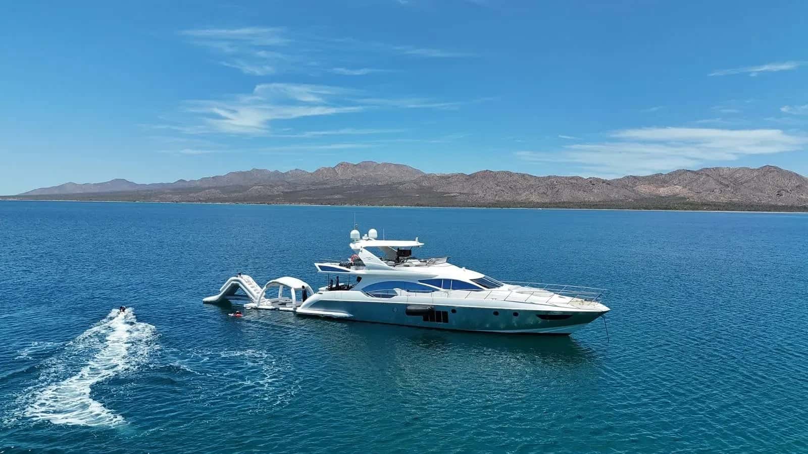 The yacht at anchor in a blue bay from the air, desert mountains along the shore