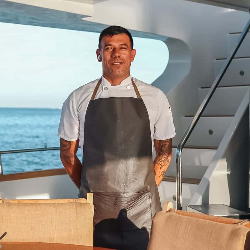 Javier in a white chef's jacket and a leather apron on a yacht's aft deck, the sea behind