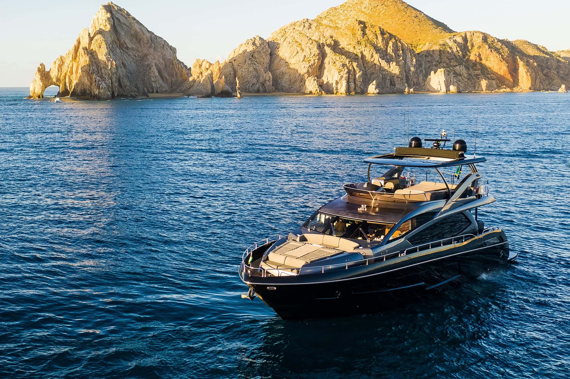 Black flybridge motor yacht on deep blue water below the rock arch and cliffs of Land's End, seen from a drone