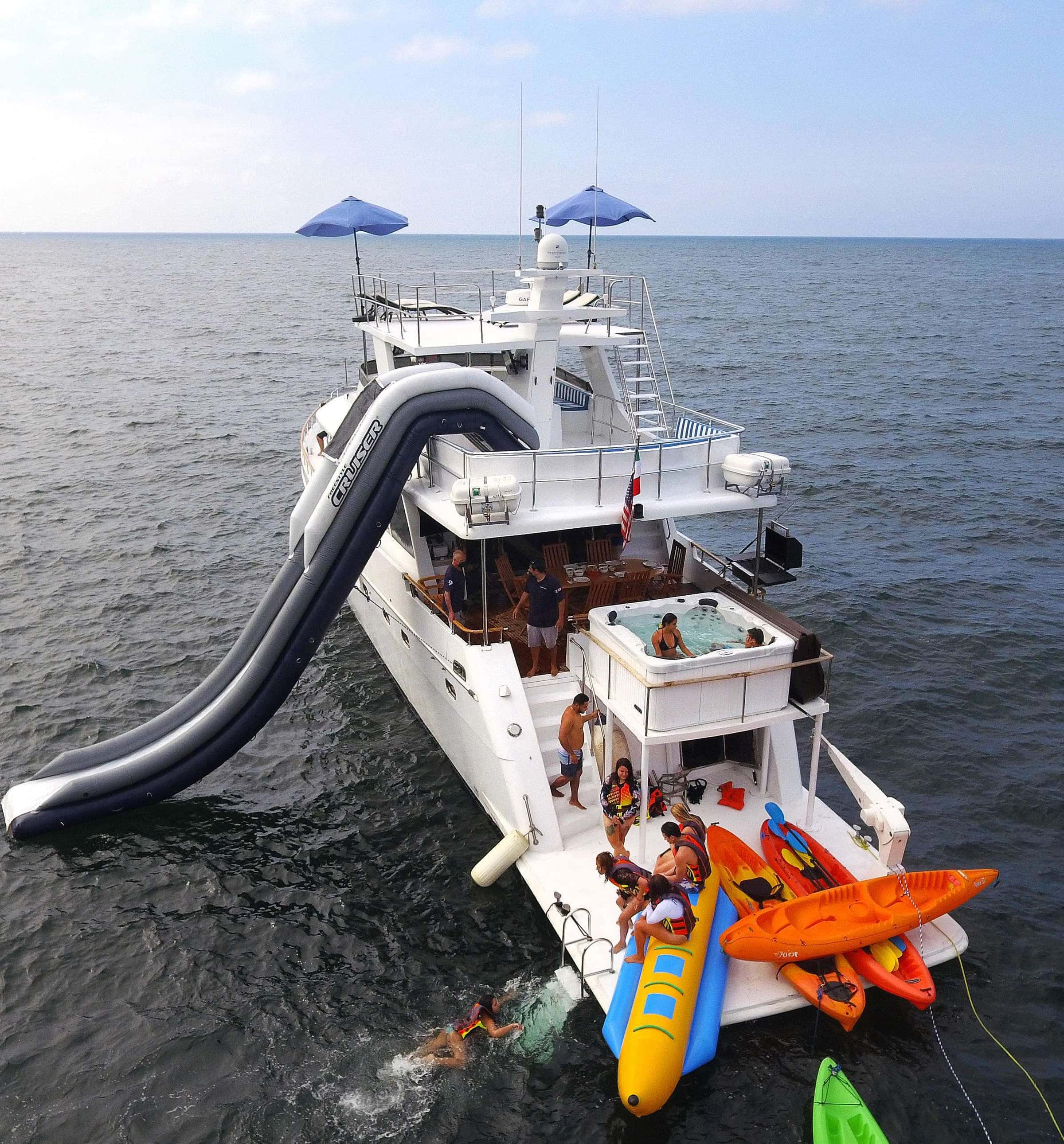 Aerial view of the stern: the inflatable slide down the side, the hot tub on the aft deck, and kayaks and a banana boat at the swim platform