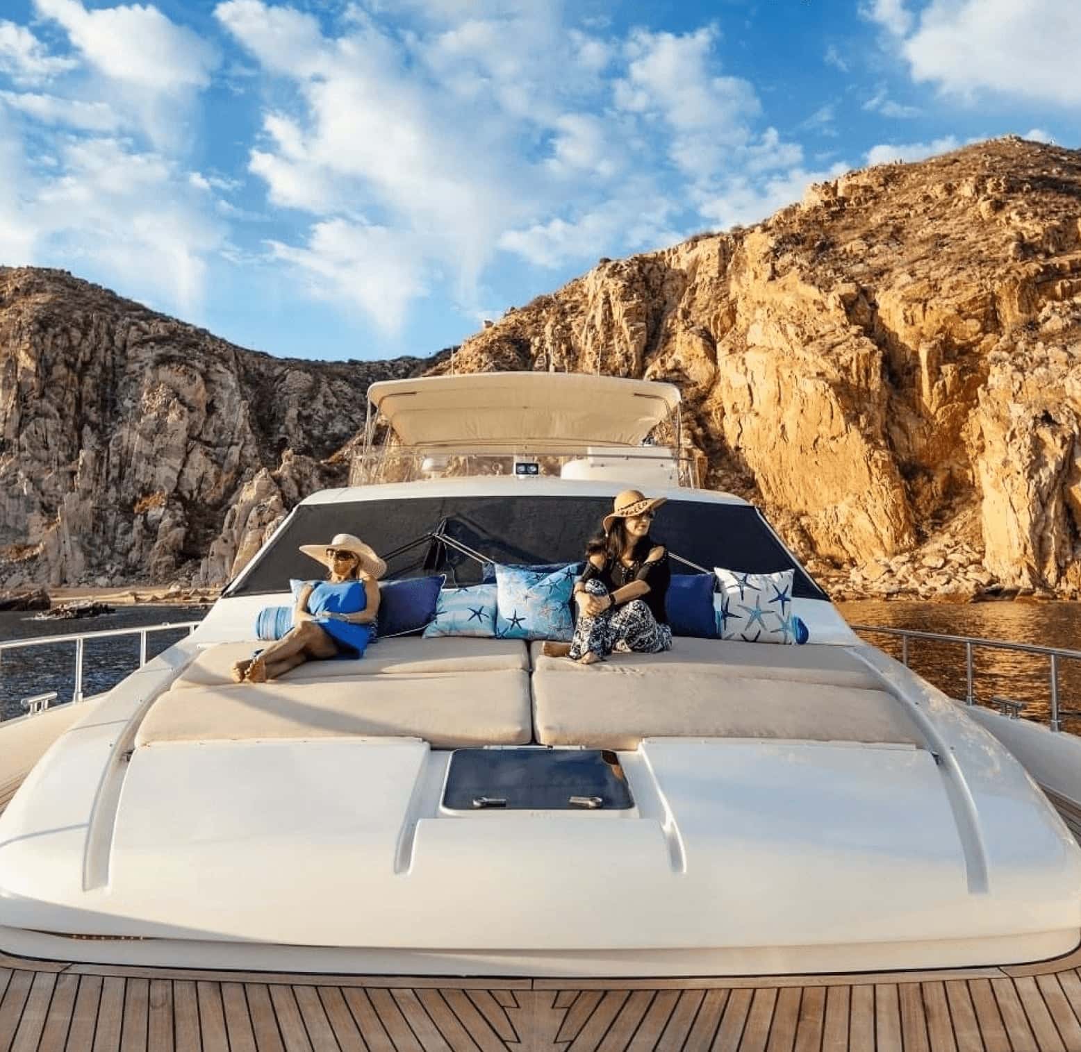 Two guests on the cushioned bow sun pad under the cliffs, the flybridge behind