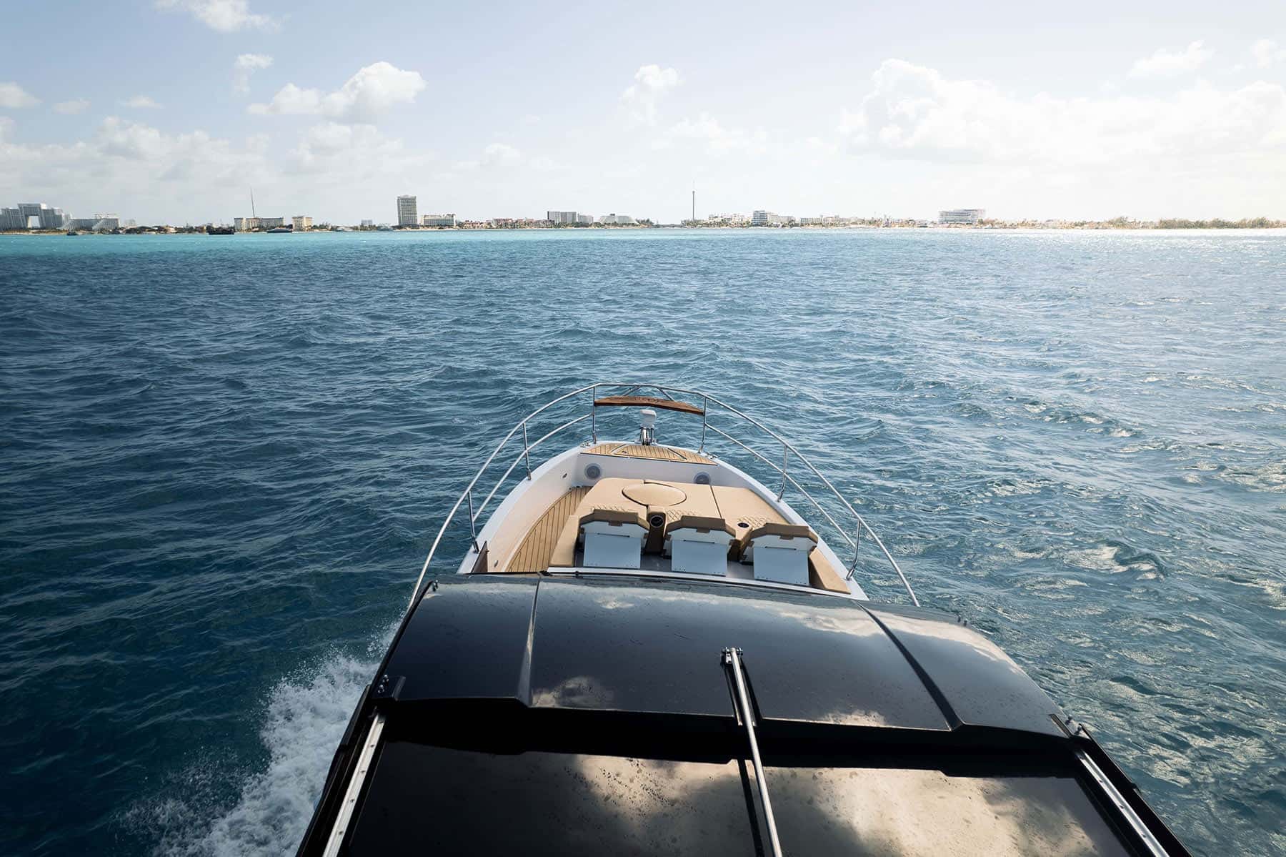 View forward over the dark hardtop to the bow sun pad and its three backrests, a hotel shoreline ahead