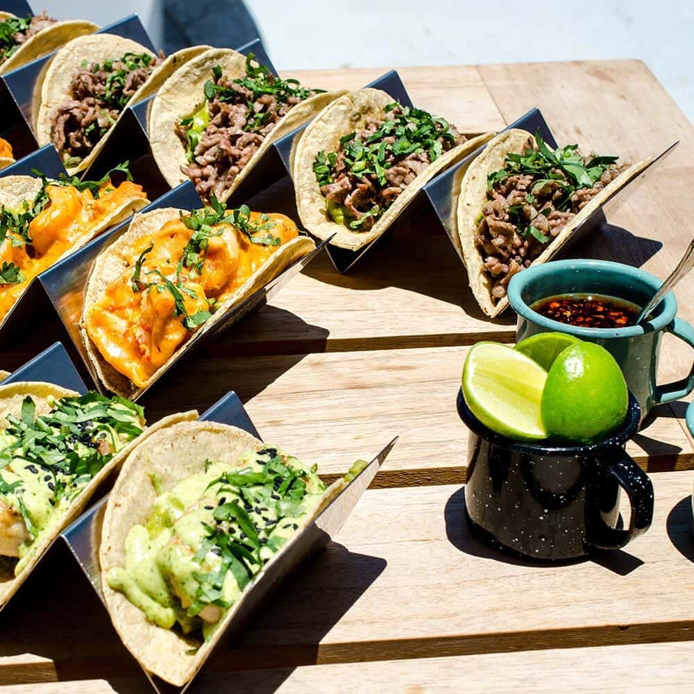 Beef, shrimp and avocado tacos in steel holders beside salsa and lime