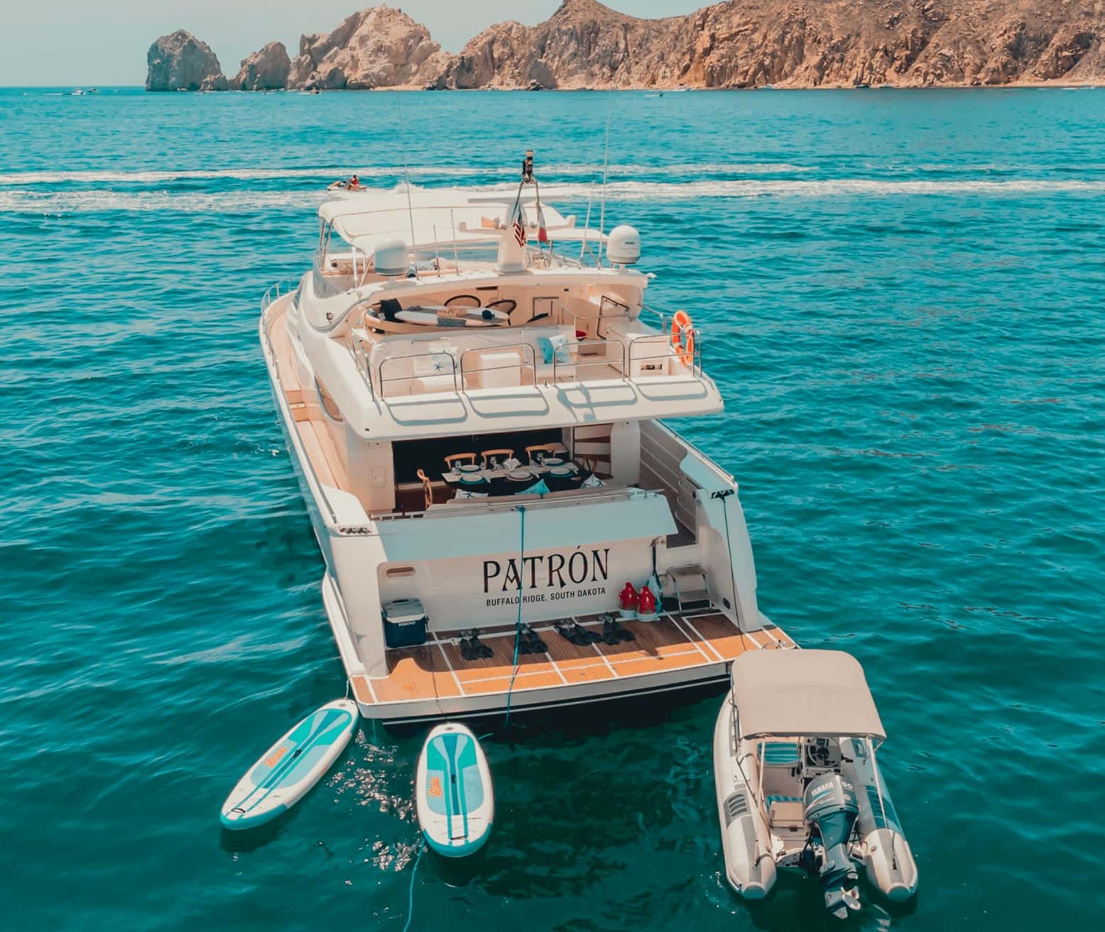 Stern of the yacht from above with a laid dining table, two paddleboards and the tender floating behind