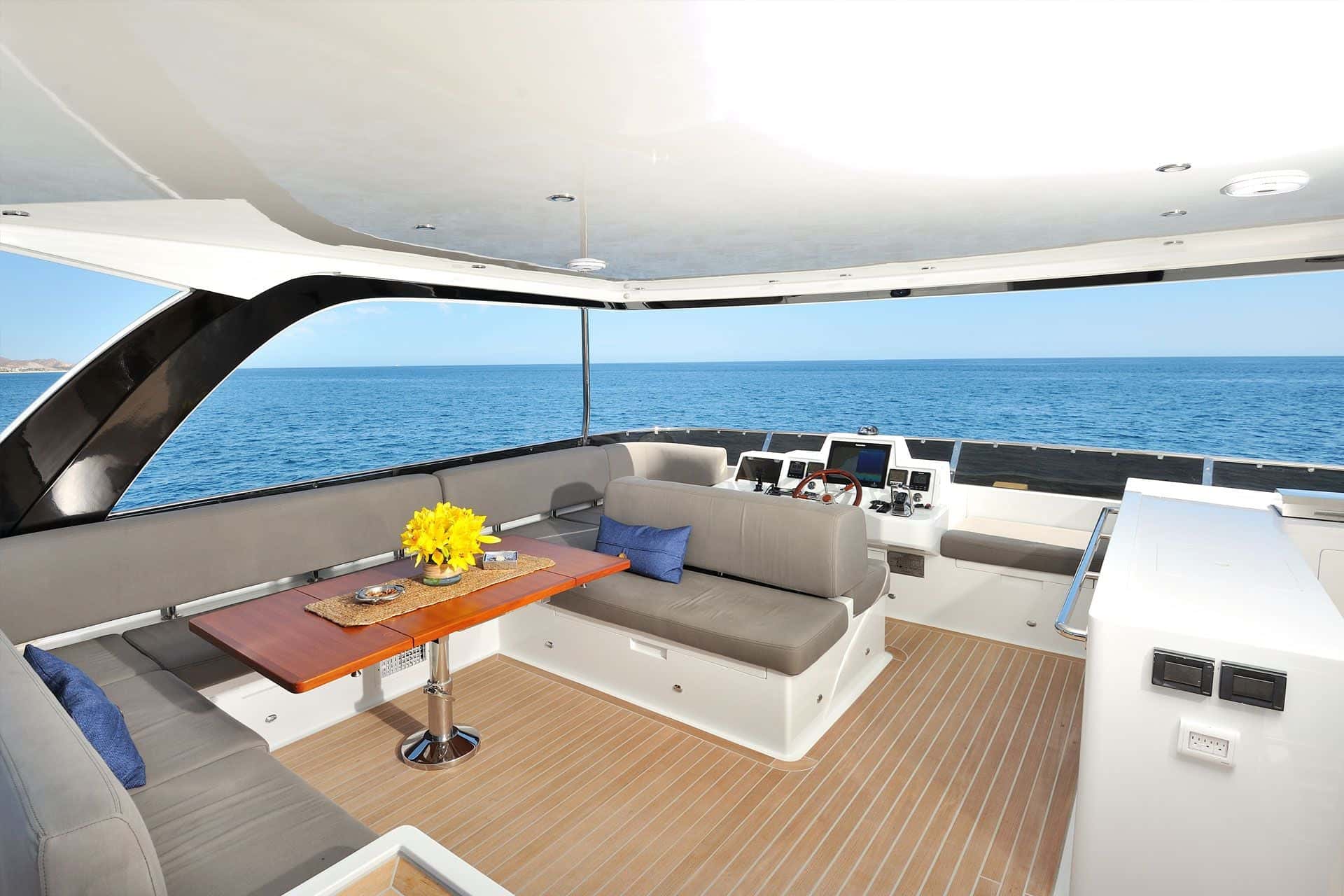 Flybridge under a hardtop with grey wraparound seating, a wooden table and the helm, open sea beyond