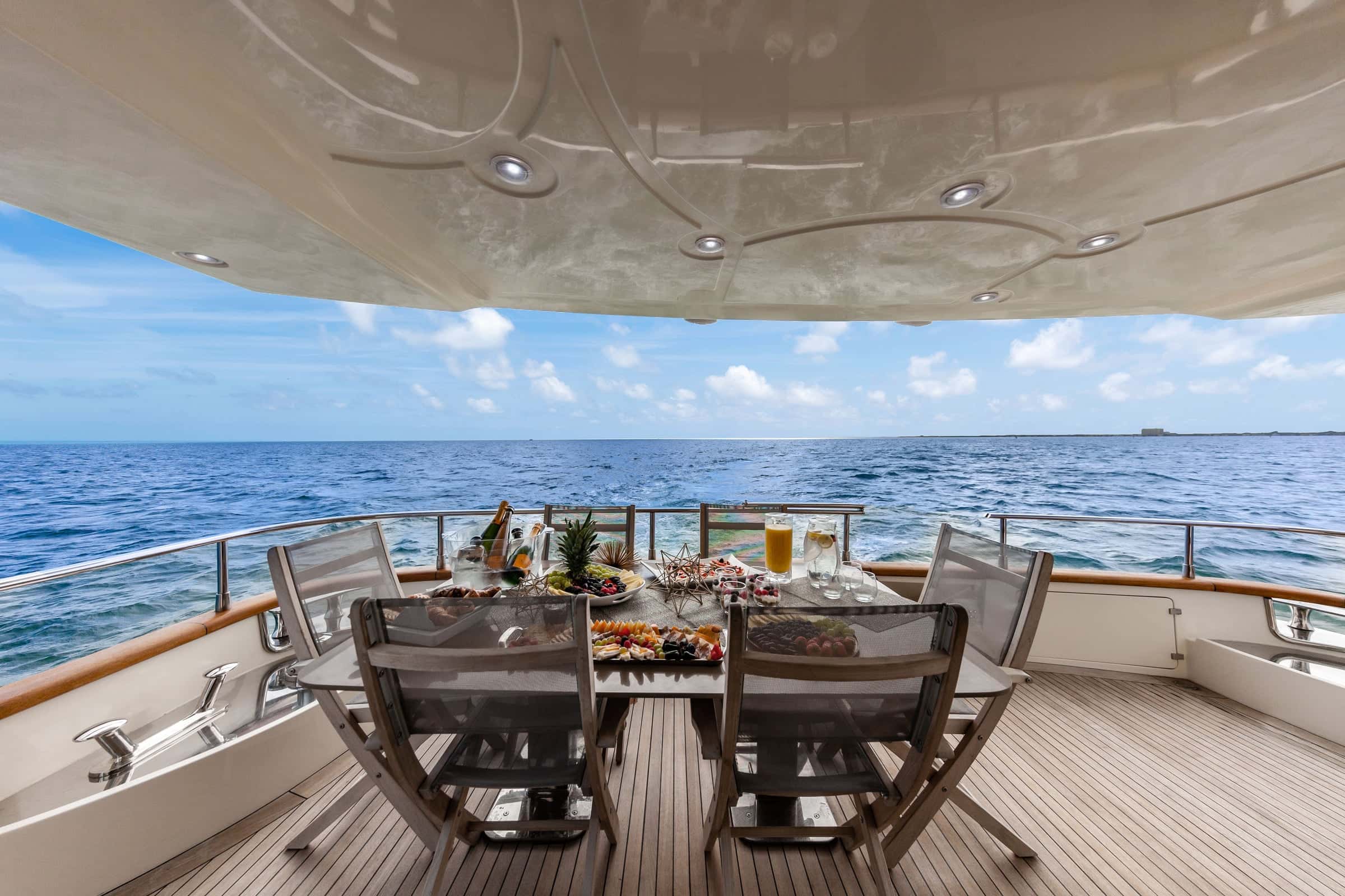 Aft deck table under the flybridge overhang, laid with fruit, platters and drinks, open sea behind